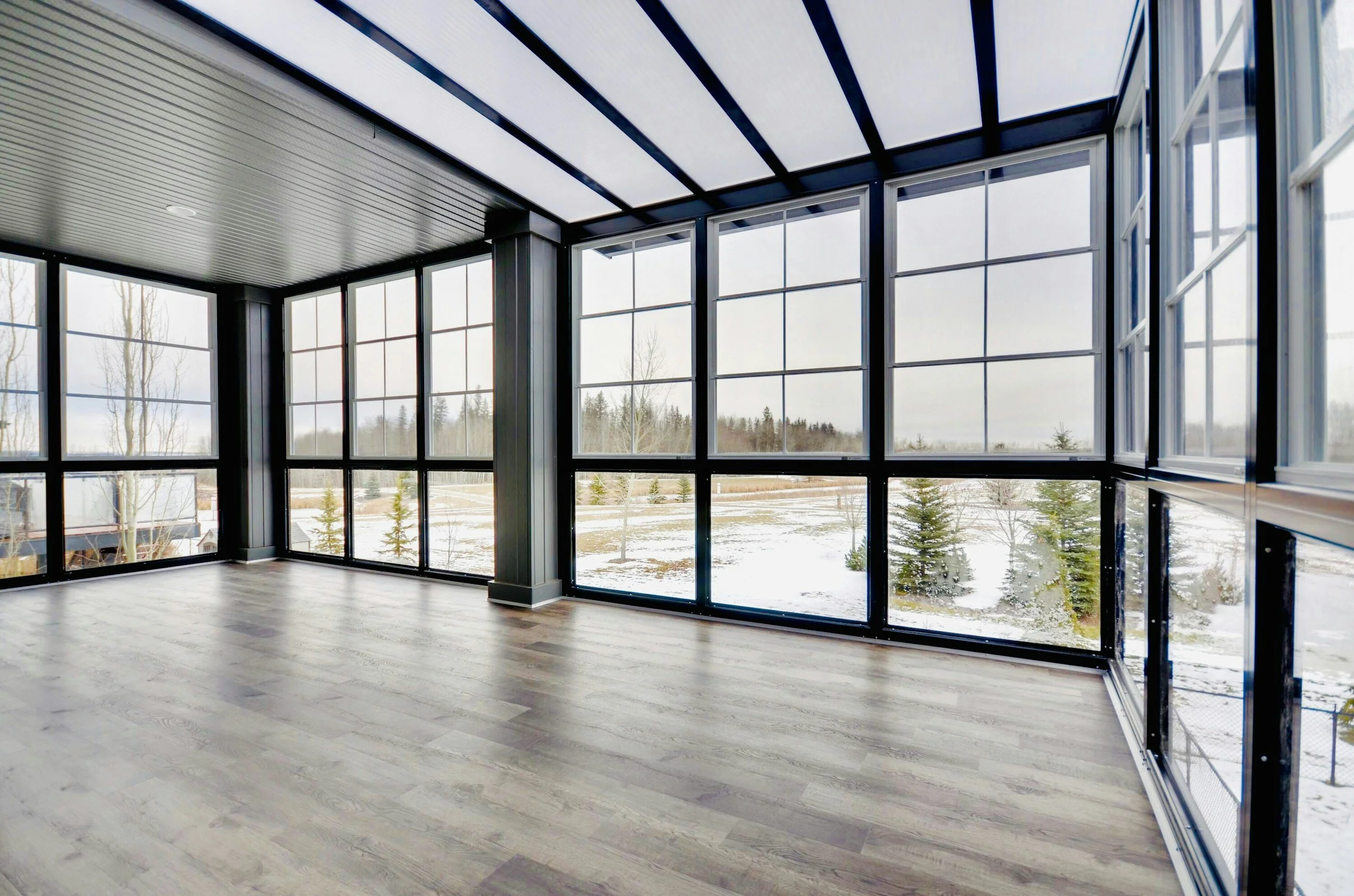 Empty sunroom with large windows showing snow-covered trees and landscape outside, wooden flooring, black ceiling beams, and a dark frame supporting the windows.