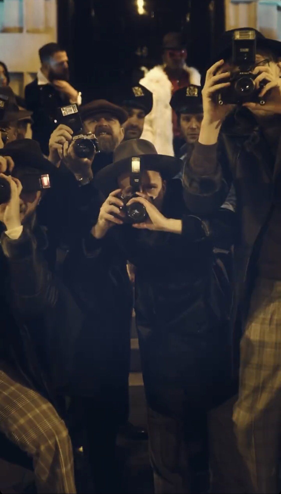 Group of photographers with cameras and flashbulbs, wearing hats and jackets, capturing photos at night in an urban setting.