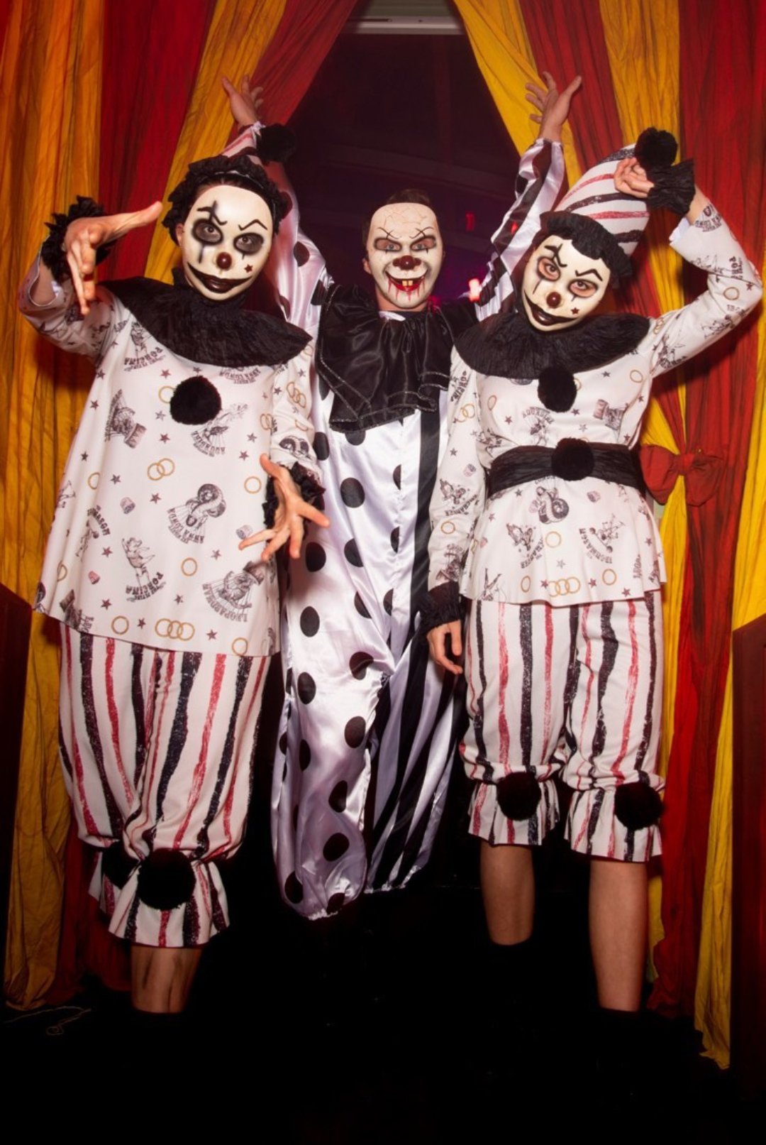Three people in eerie clown costumes and makeup posing in front of colorful drapes.