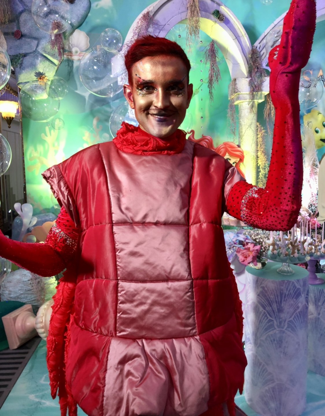 Person in a red crab costume with vibrant background featuring sea-themed decor and bubbles.