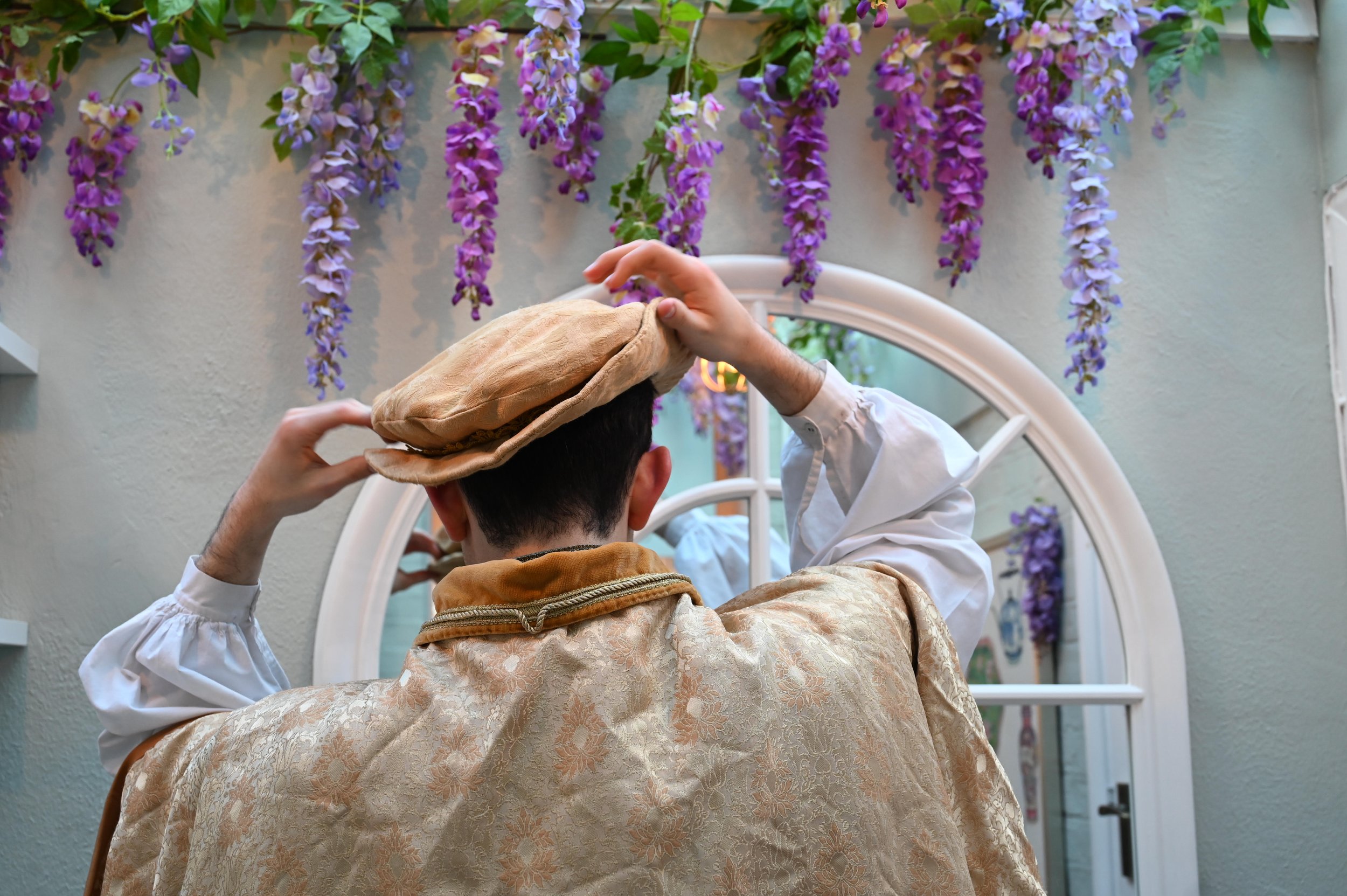 Person in vintage costume adjusting hat in front of a mirror with hanging purple flowers above.