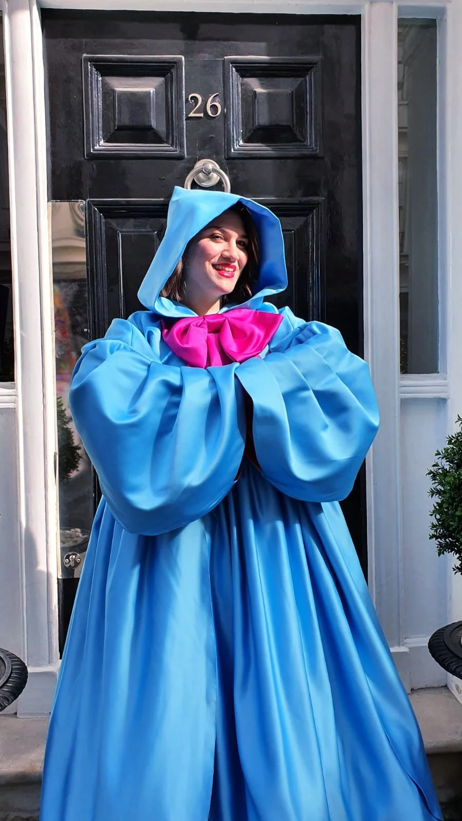 A woman dressed as a fairy tale character, wearing a bright blue hooded cape with puffy sleeves and a large pink bow around her neck, smiling in front of a black door with the number 26.