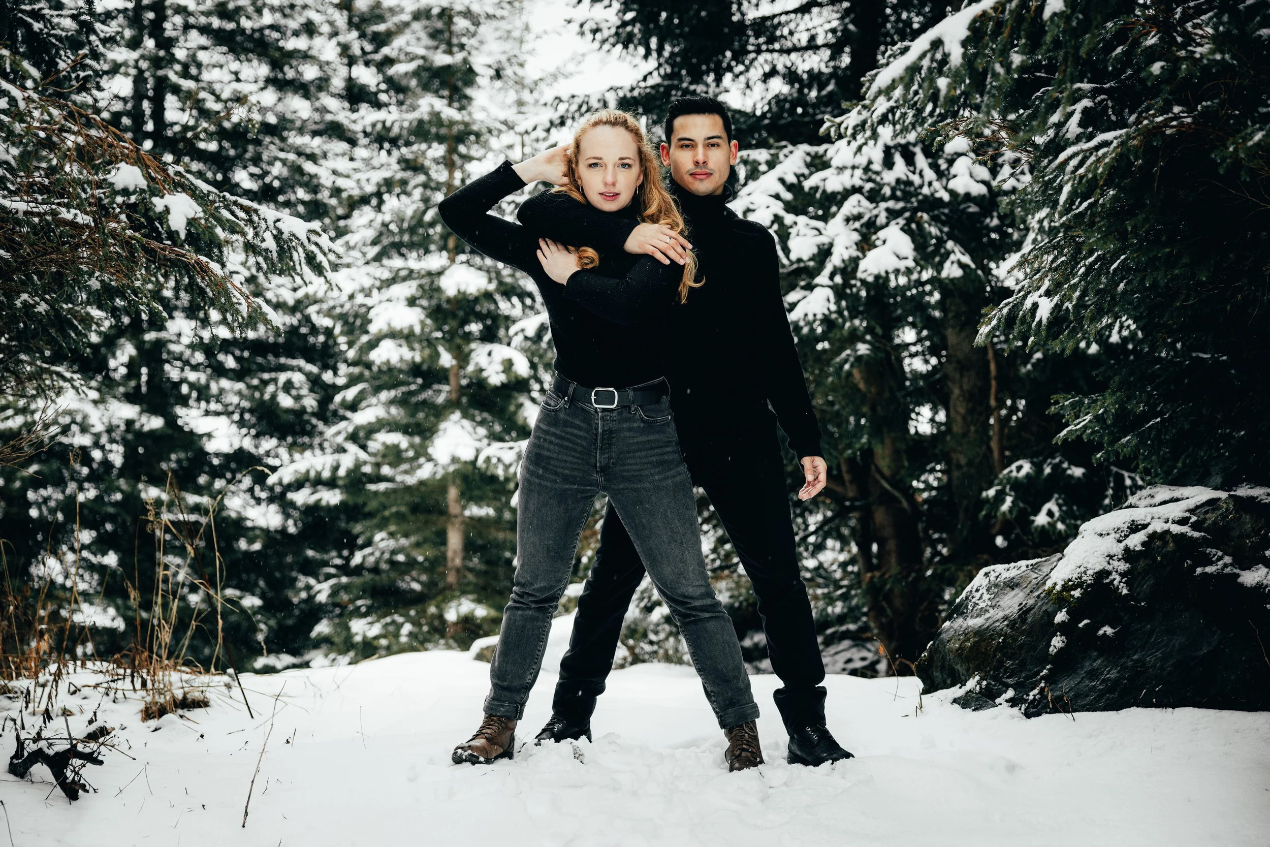 Un couple posant dans une forêt enneigée, vêtus de noir.