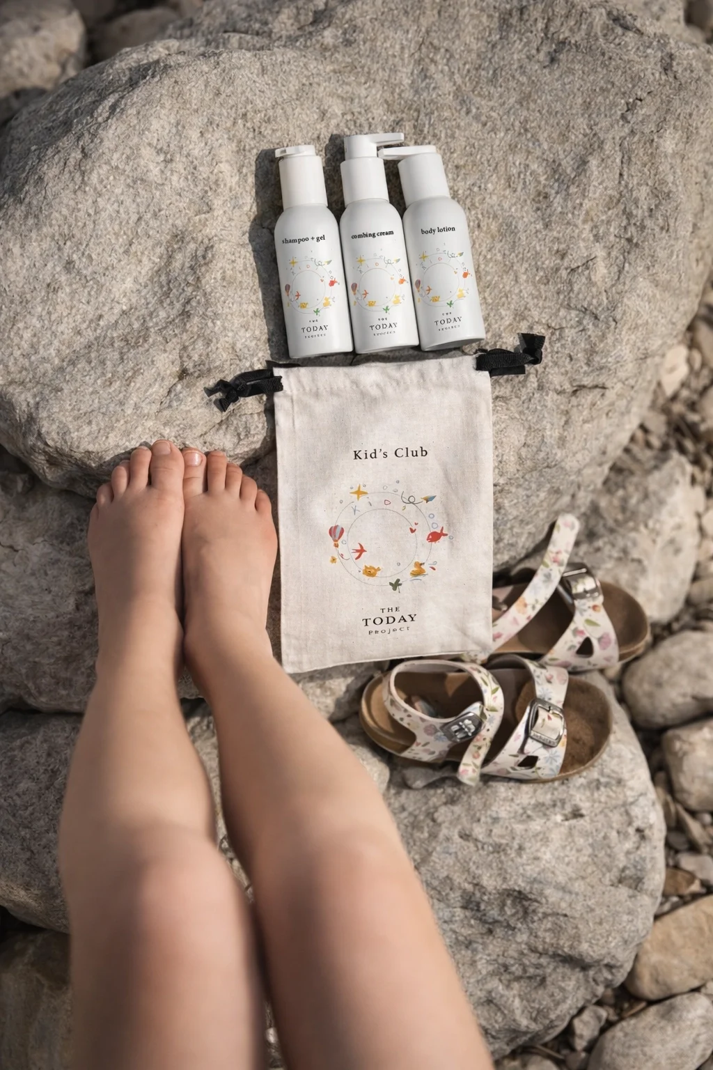 A pair of small feet with toes on a rocky surface, with three white bottles of lotion or soap labeled 'shampoo + gel,' 'coming cream,' and 'body lotion,' a cloth bag with 'Kid's Club' and 'THE TODAY project' printed on it, a pair of sandals with floral straps, and a large rock in the background.