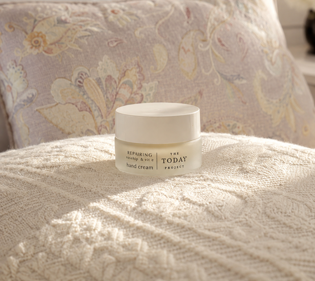 Ein Tiegel Handcreme mit der Aufschrift „Repairing Rosehip & Vit E Handcreme“ von The Today Project, platziert auf einer strukturierten beigen Decke, im Hintergrund ein Kissen mit Blumenmuster.
