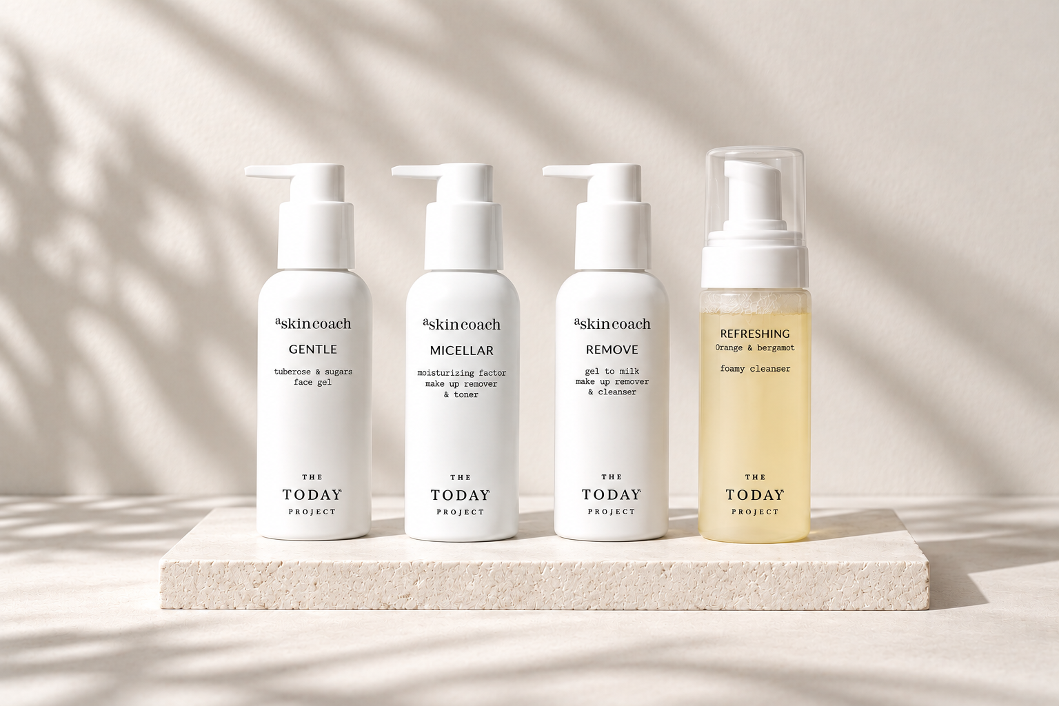 Cuatro frascos de productos para el cuidado de la piel sobre una base blanca con textura, etiquetados como «gel facial», «desmaquillante y tónico», «desmaquillante y limpiador» y «limpiador espumoso», con sombras naturales sobre un fondo claro.