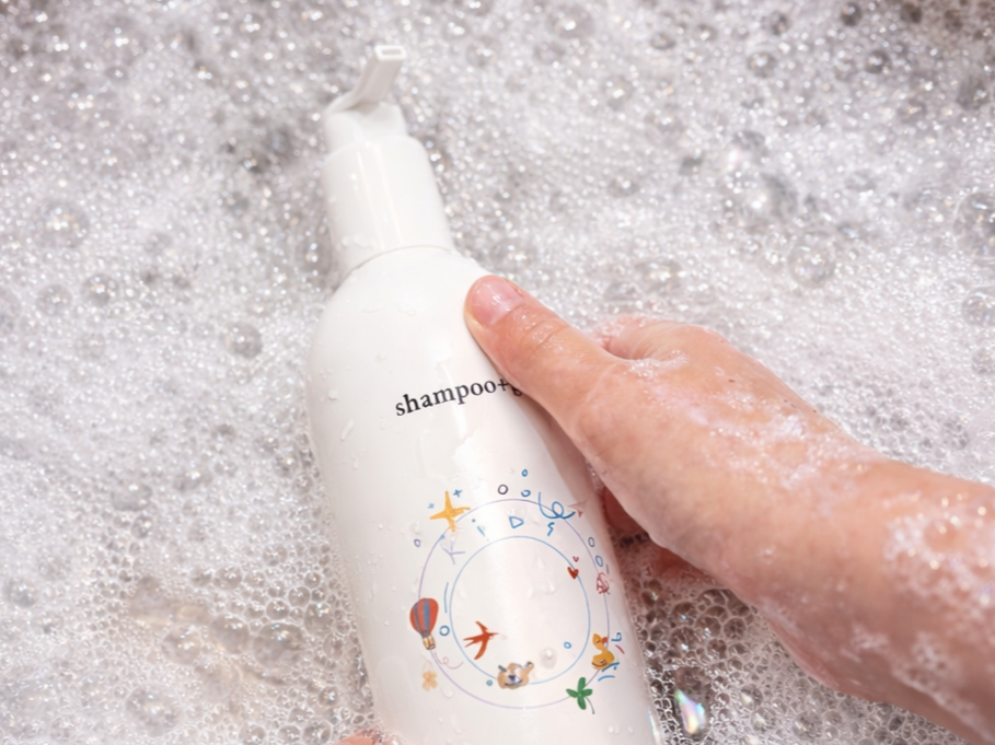 A person holding a white shampoo bottle with a decorative label, surrounded by soap bubbles in a bath or shower setting.