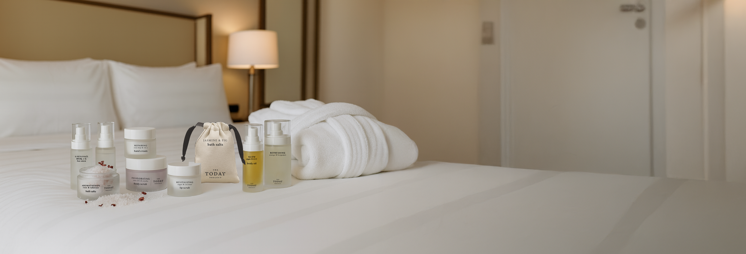 Cama de hotel con sábanas y toallas blancas, con productos para el cuidado de la piel y el baño dispuestos sobre ella, cerca de una lámpara y una pared beige.