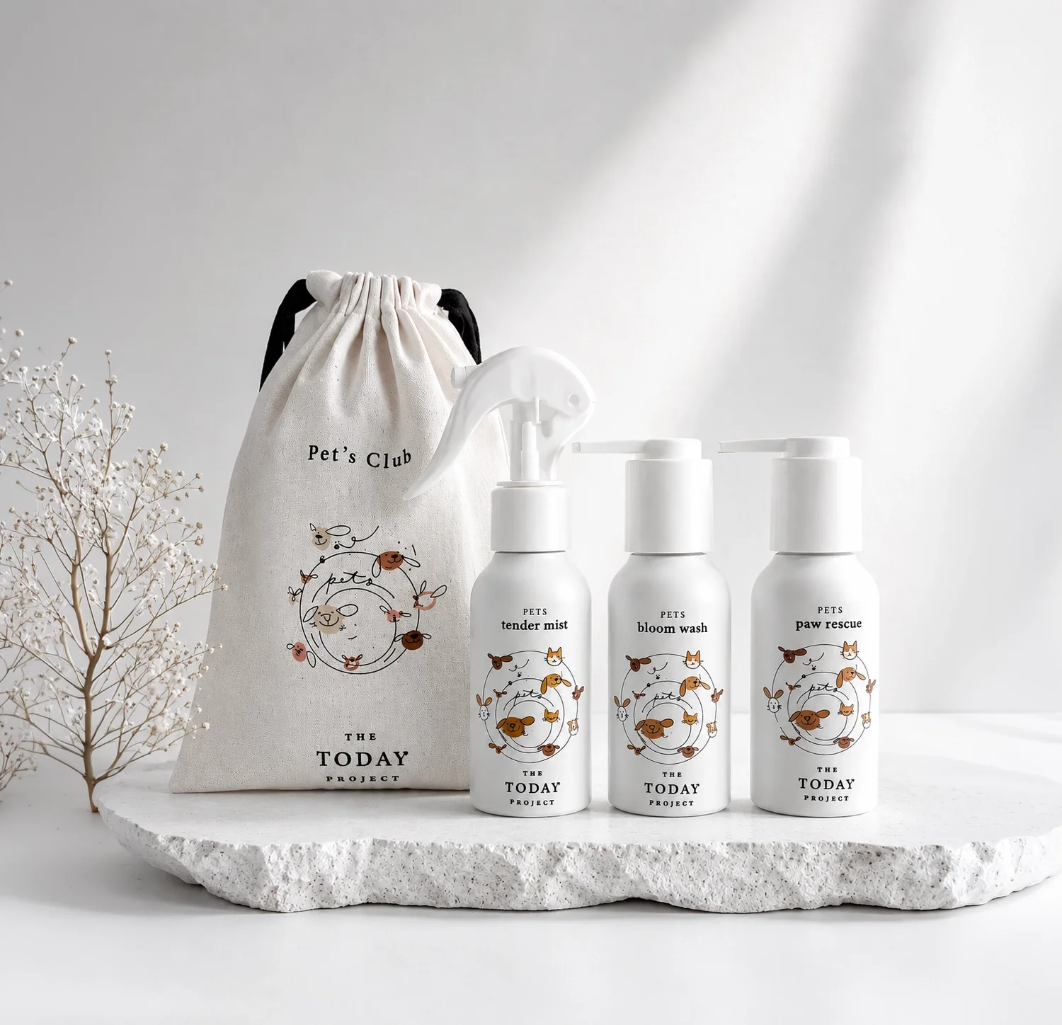 A set of pet grooming products, including three white spray bottles labeled as tender mist, bloom wash, and paw rescue, with a matching cloth bag and a small dried plant on a textured white surface against a plain background.