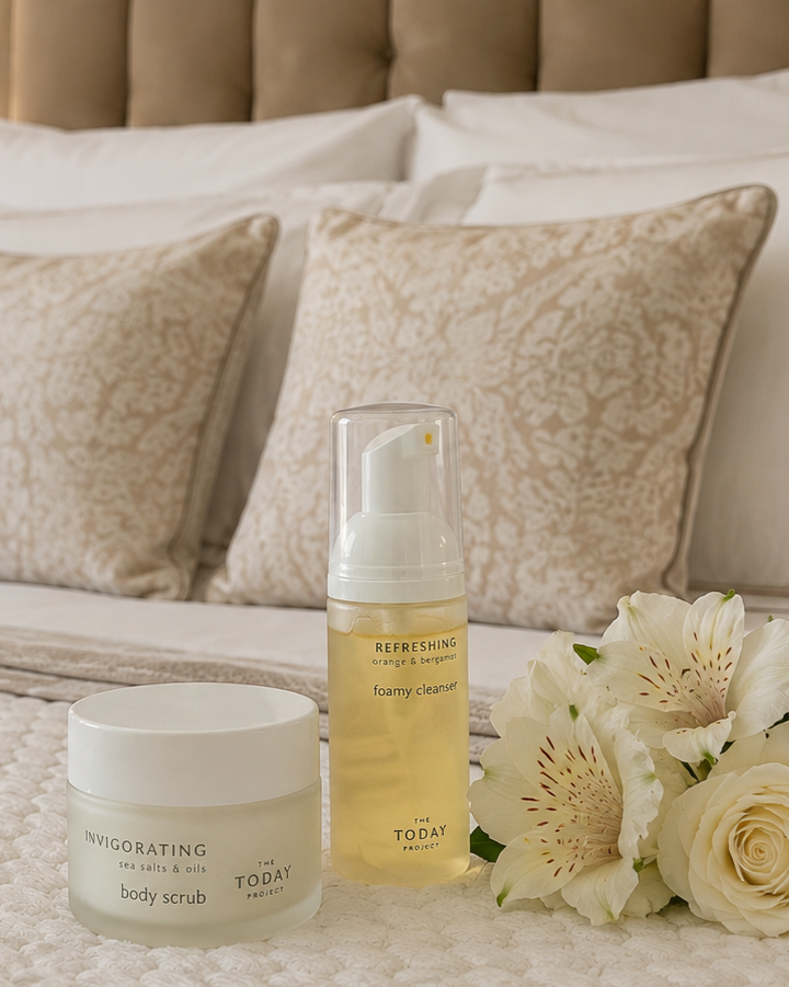 Hautpflegeprodukte, darunter ein Körperpeeling, ein schäumendes Reinigungsmittel und Blumen auf einem Bett mit beigen Kissen.