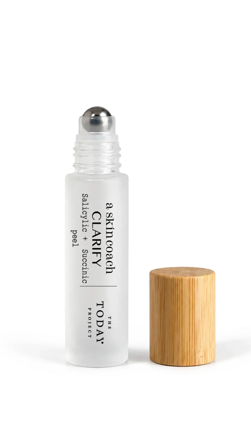Un producto para el cuidado de la piel en un frasco blanco con un aplicador de bola metálico, etiquetado como «Skin Coach C-Larity», un sérum con ácido salicílico y peeling succínico, junto a un tapón de madera y una superficie blanca con una gota de sérum.