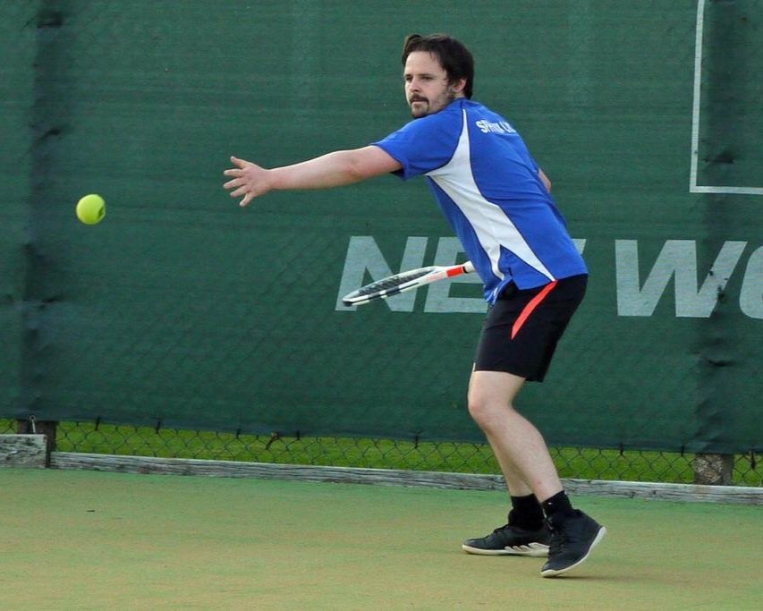 A young man wearing glasses, a navy hoodie, and blue Adidas sweatpants playing tennis on an outdoor court during autumn.
