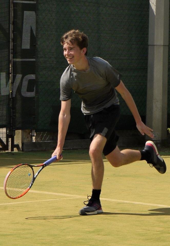 Young woman in sporty blue top and shorts standing on tennis court, holding tennis racket, in a stretching pose with one leg extended parallel to the ground.