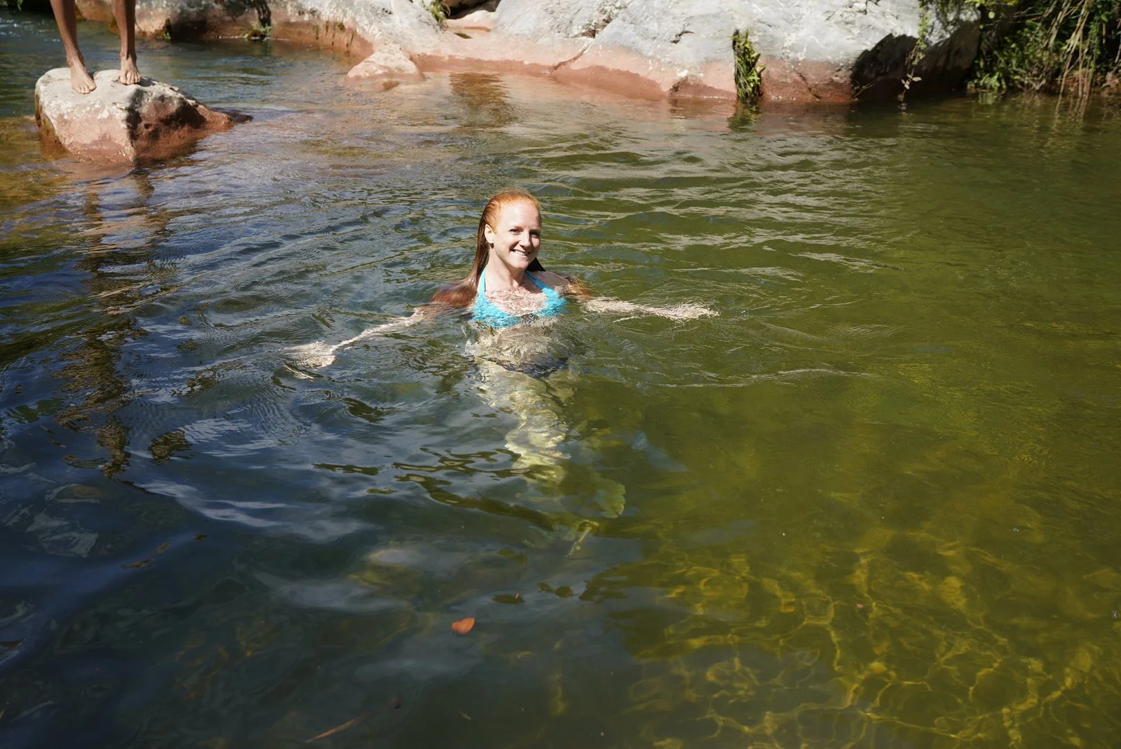 Una mujer sonriente nadando en un río con agua de color verde y personas de pie en las rocas en la orilla.