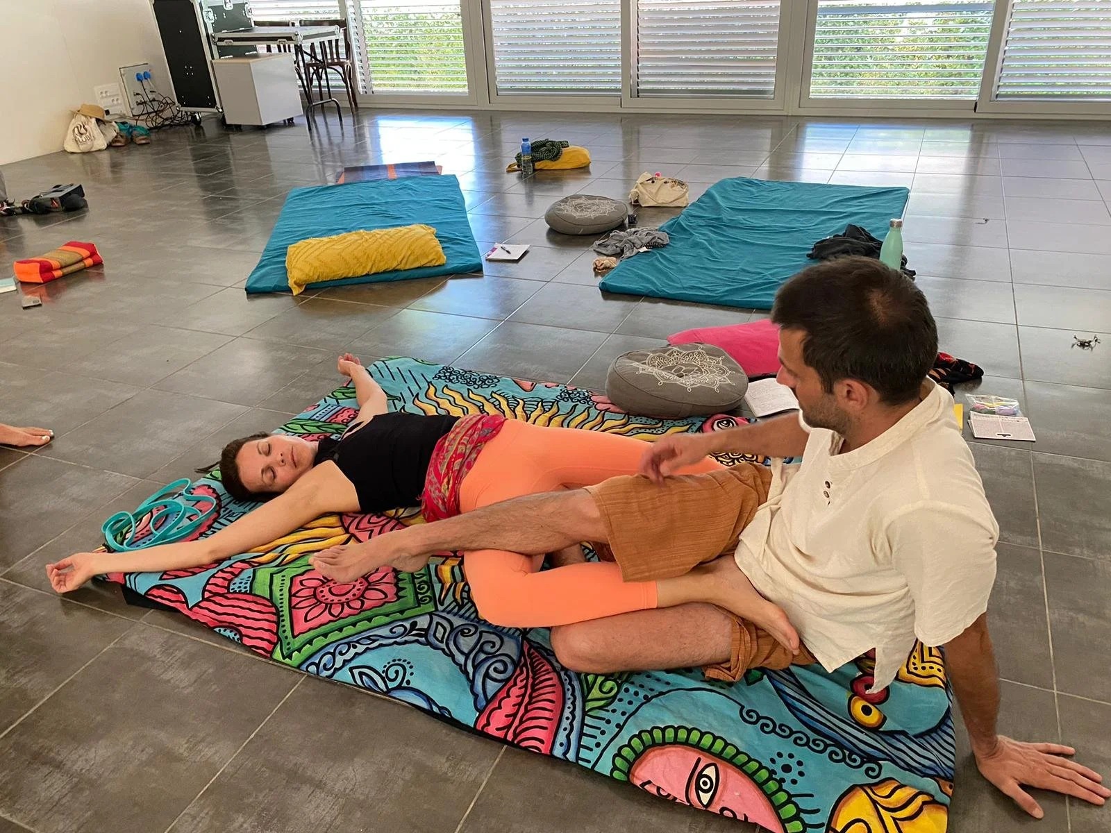 Una mujer acostada en una postura de yoga con un hombre que le ayuda a estirar las piernas, en un espacio interior con muchas almohadas y colchonetas en el piso.