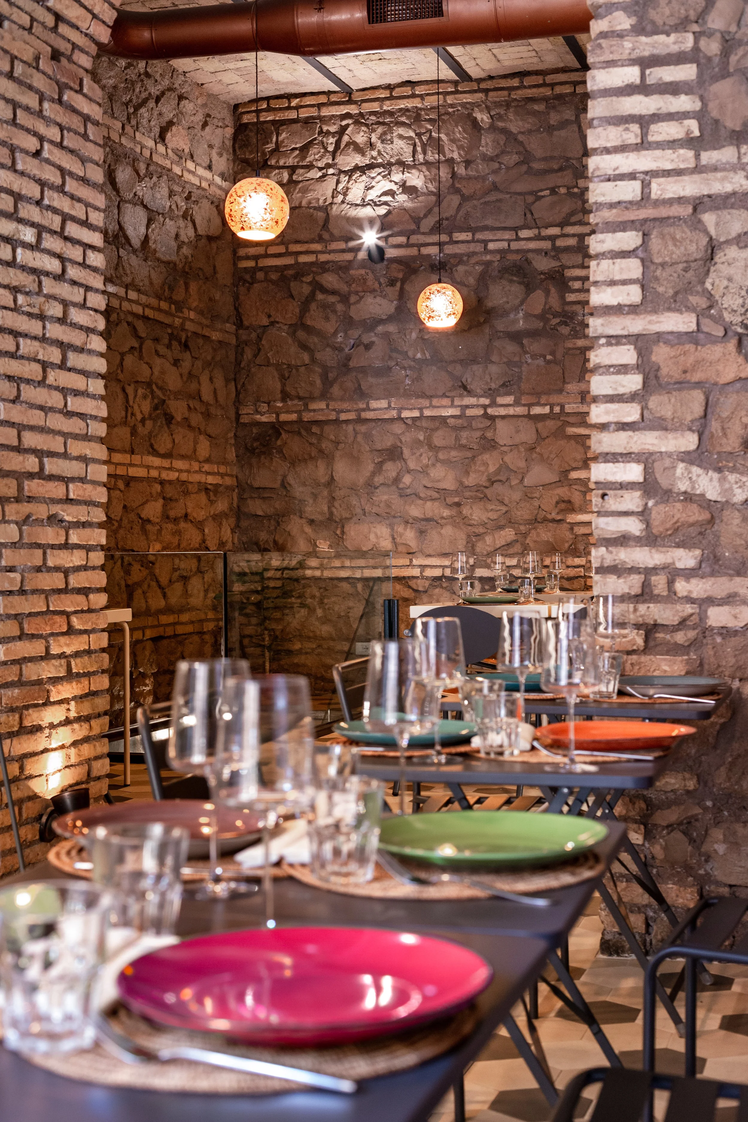 Interno di un ristorante con tavoli apparecchiati, piatti colorati e bicchieri, illuminazione moderna e pareti in pietra a vista.