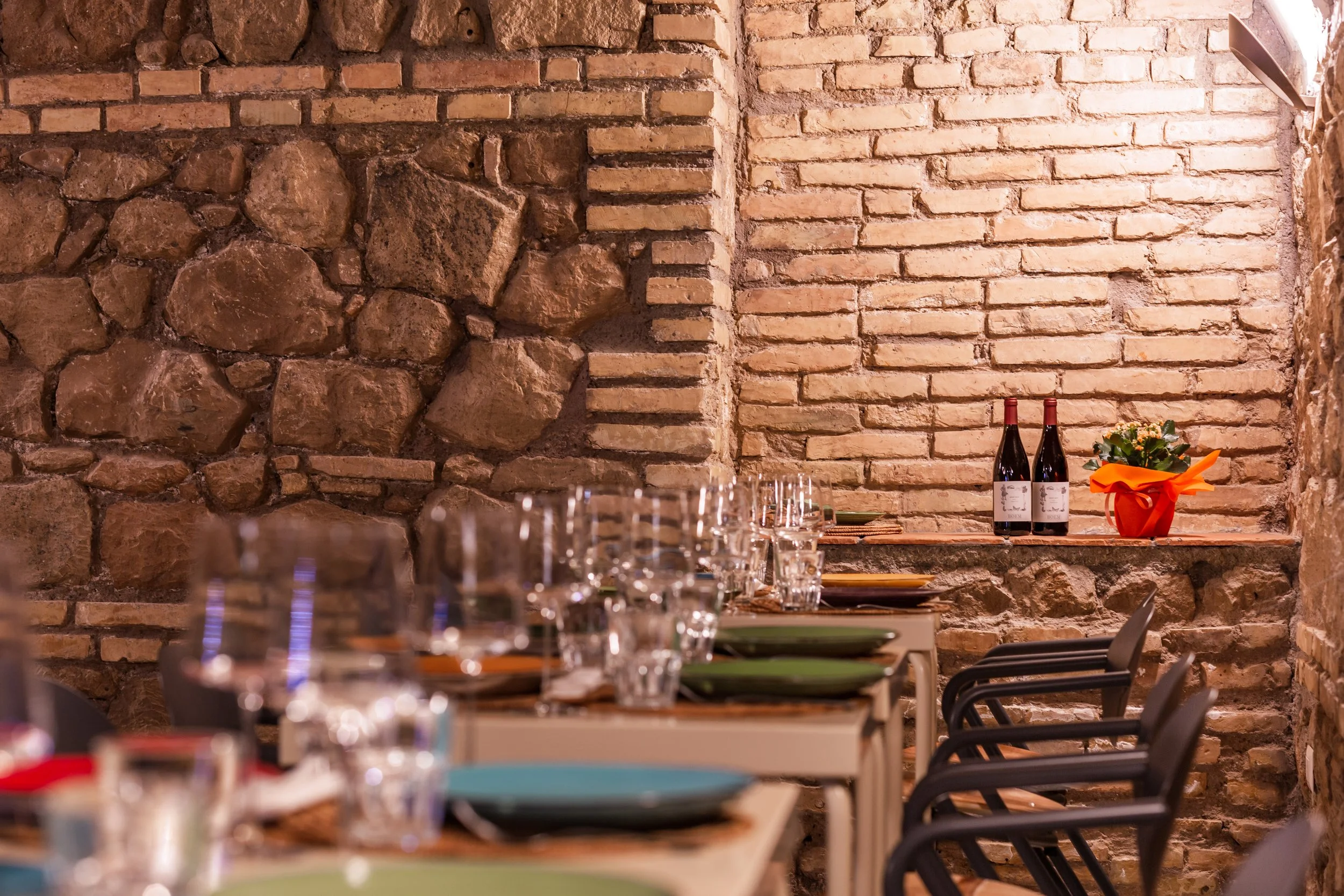 Interno di un ristorante con pareti in mattoni a vista, tavoli apparecchiati con bicchieri, piatti e posate, due bottiglie di vino e una pianta in vaso con fiori gialli su una mensola.
