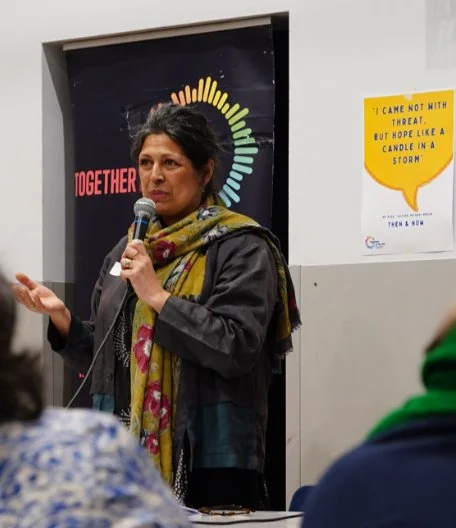 Sita Brahmachari speaking into a microphone with people in the foreground
