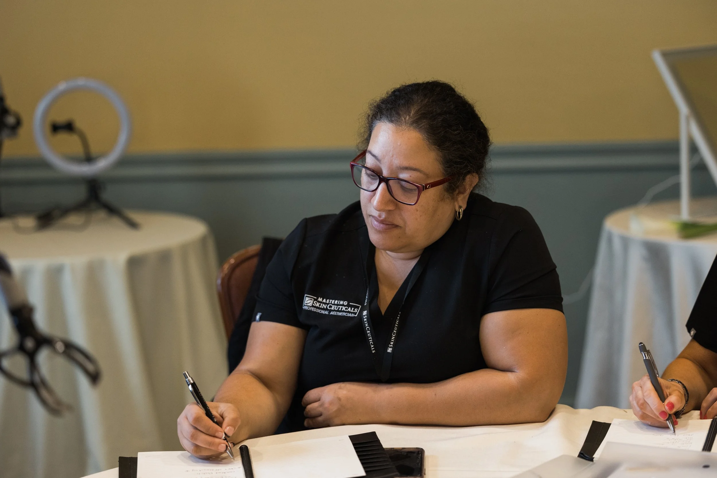 A woman with glasses and a black shirt sitting at a table, writing on a notepad.