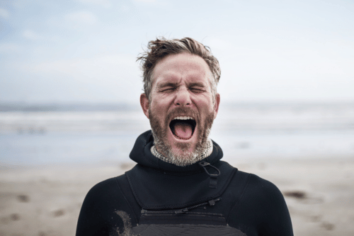 A white man in a wetsuit appears to shout at the camera (or no one in particular) with the sea in the background.