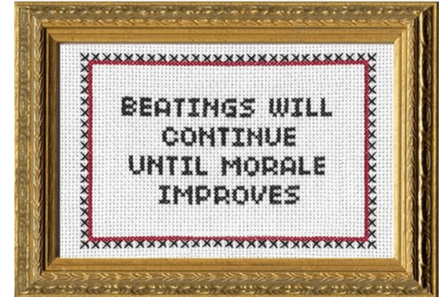 A picture frame containing cross-stitched text reading "Beatings Will Continue Until Morale Improves"