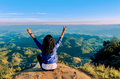 A woman sits on a cliff, facing away from the camera and flashing peace/victory signs with both arms held high.