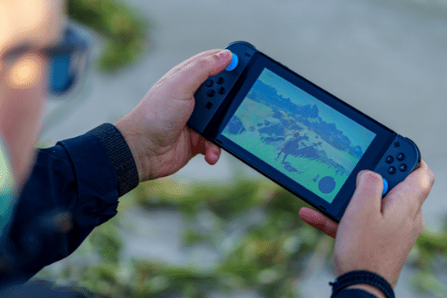 One of my favorite forms of procrastination! Photo of adult hands holding a Nintendo Switch and playing Legend of Zelda: Breath of the Wild. The person's head is mostly out of frame and out of focus but is wearing glasses.