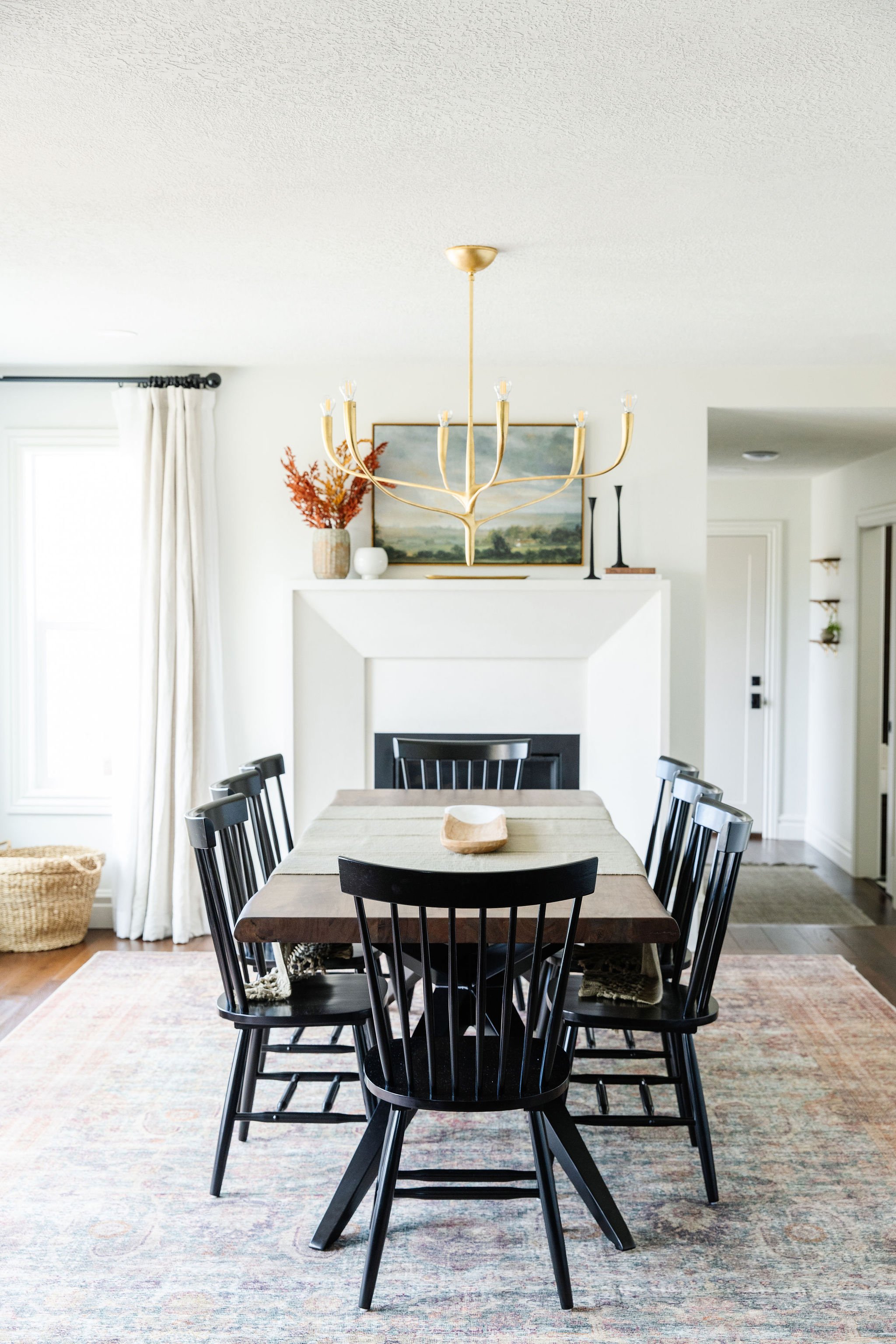  Liz Powell designs an inviting dining area with an elongated dining table positioned on an area rug with a golden chandelier centered above. Fireplace golden chandelier black chairs wooden table #beveledfireplace #largediningtable #diningroomarearug