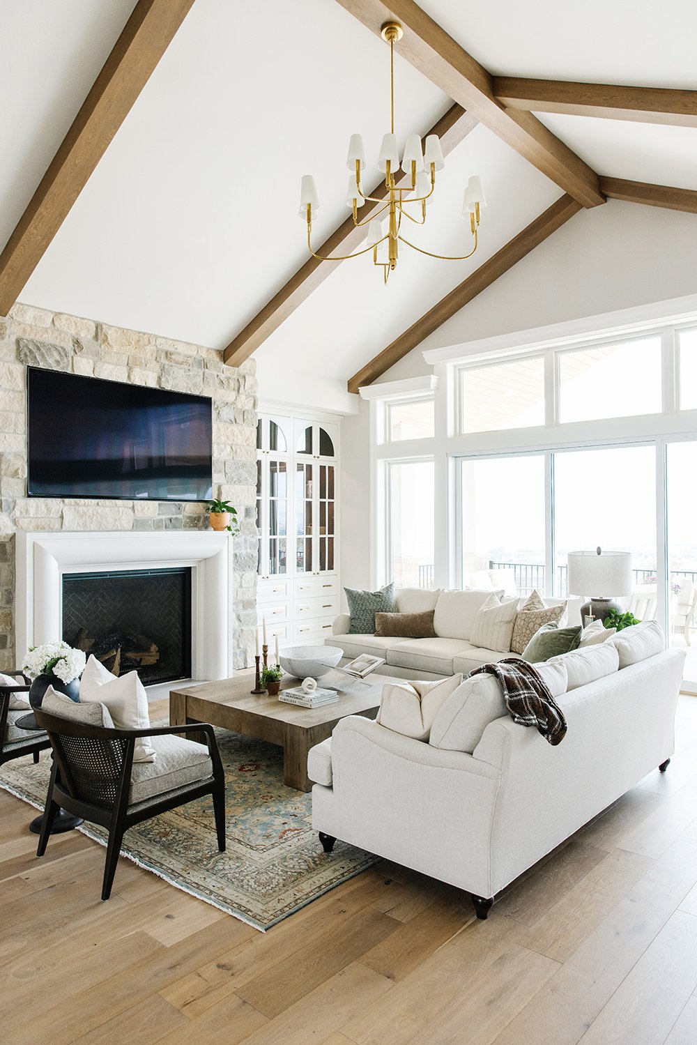  The exposed beams focus on the high ceilings and full windows. The light coloring of the rock on the fireplace flows with the neutral color palette. Exposed beams vaulted ceilings stone fireplace #lizpowell #exposedbeams #goldenchandelier 