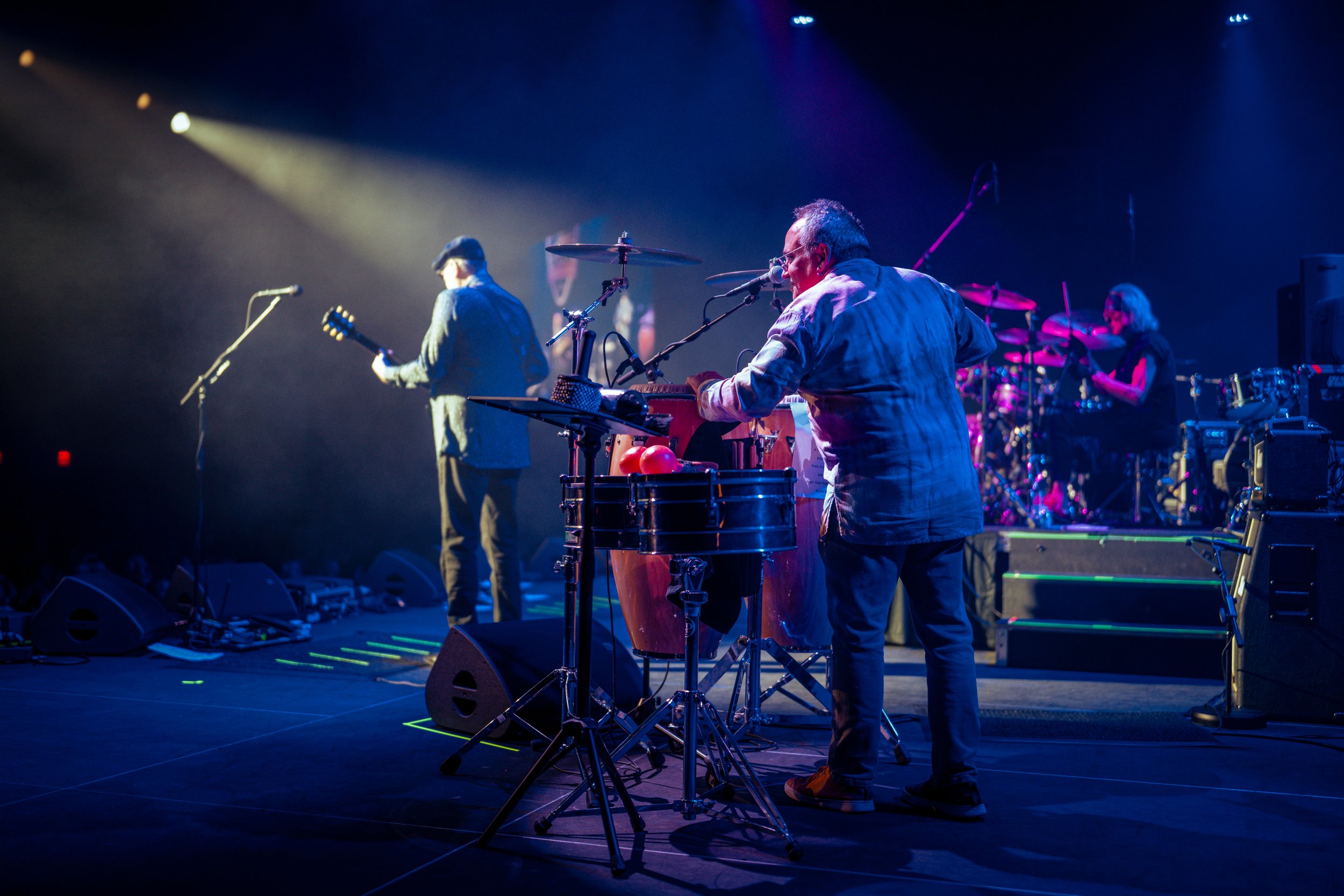 Musicians performing on stage with various instruments, including drums, a guitar, and electronic equipment, under colorful stage lighting.