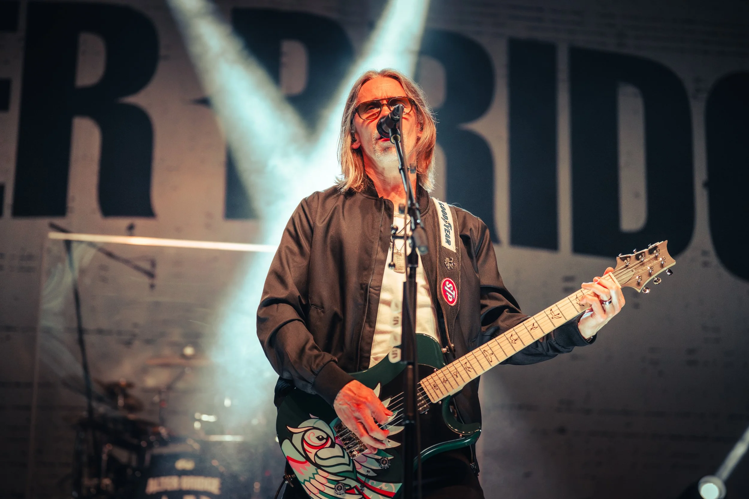 A male musician with long hair, glasses, and a beard playing an electric guitar on stage under bright lights.