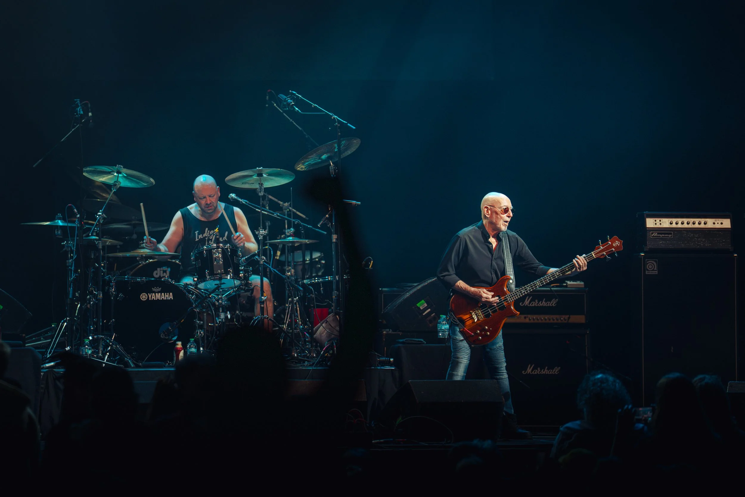 Two musicians performing on stage: one playing drums at the left and another playing electric bass guitar at the right, with amplifiers and microphones around them.