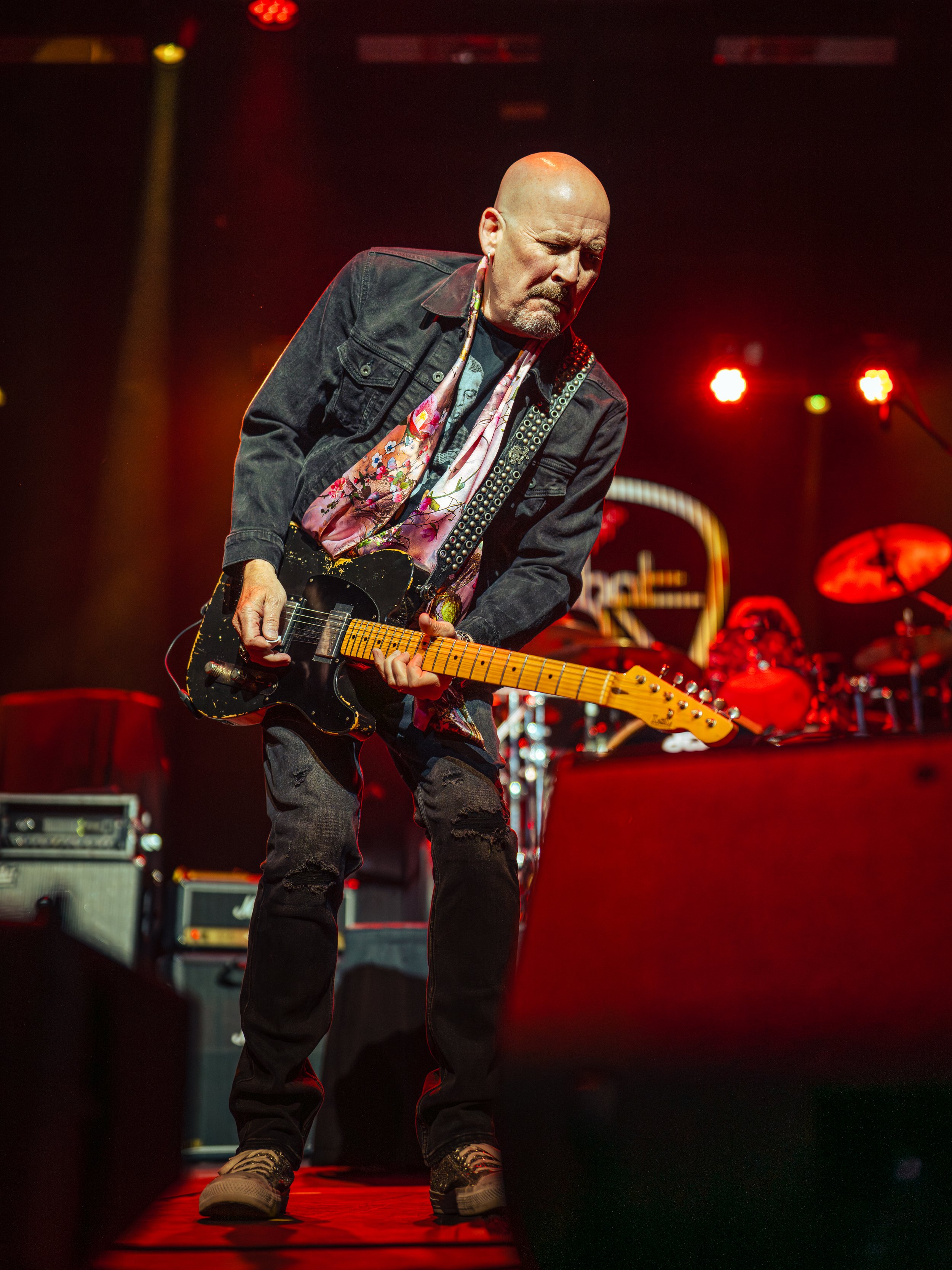 A musician performs on stage, playing an electric guitar while wearing a black denim jacket, a graphic t-shirt, ripped jeans, and boots, with colorful scarves around his neck; stage lights illuminate the scene.