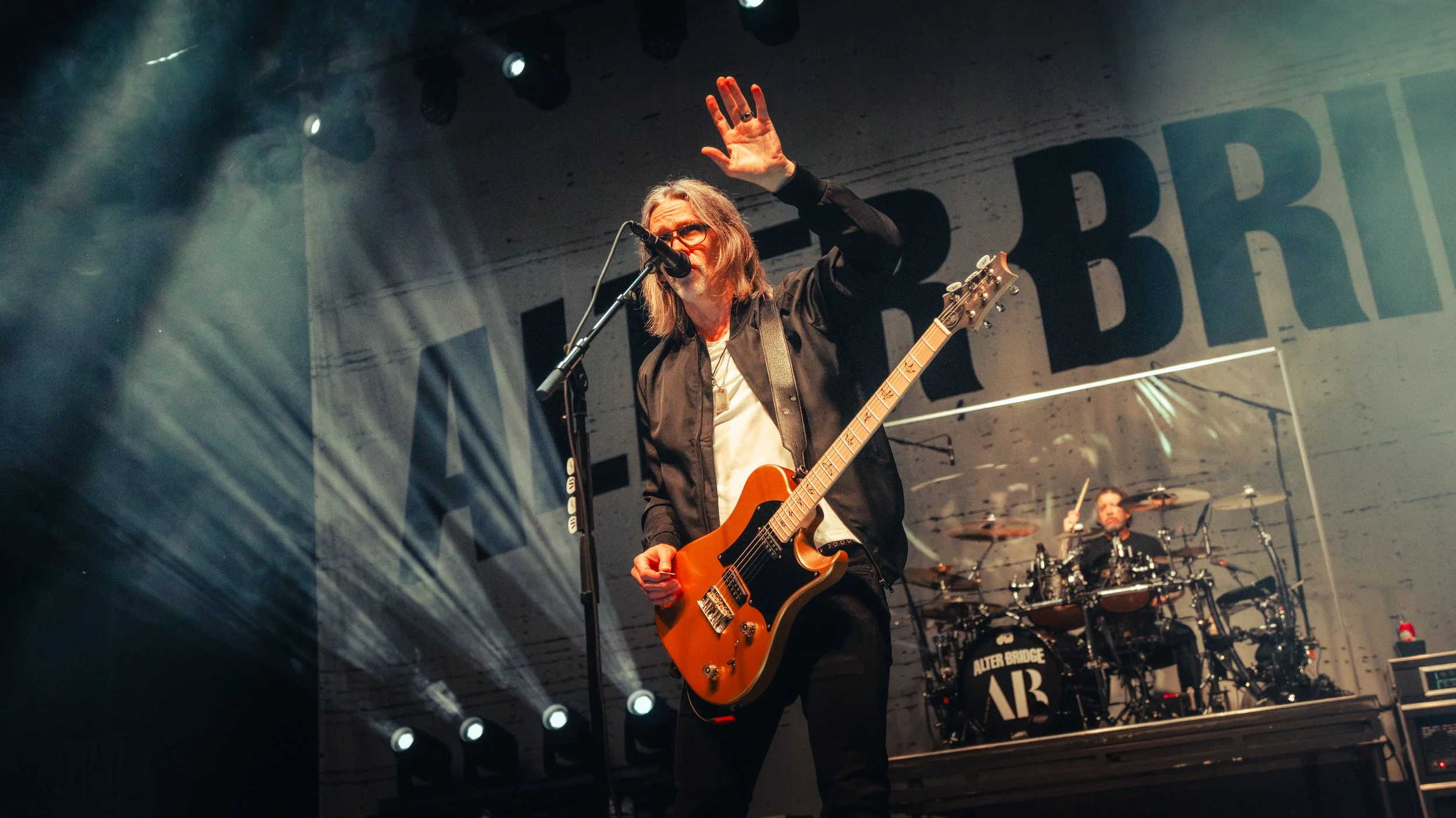 A musician with long hair, glasses, and a black jacket plays an orange electric guitar on stage, waving to the audience with a raised hand, with a drummer visible in the background and stage lights illuminating the scene.