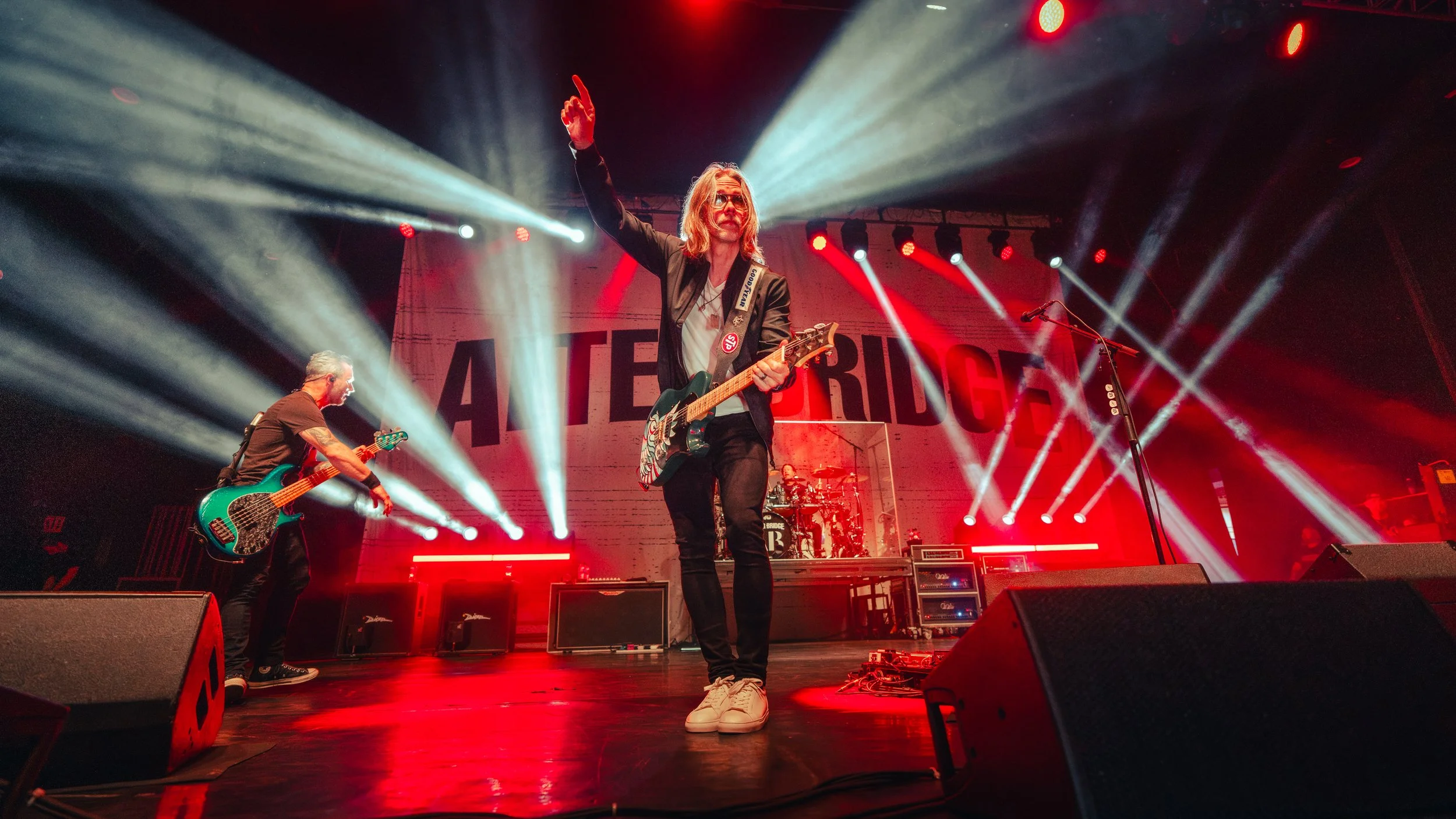 A band performing live on stage with dramatic lighting. The lead singer plays guitar and raises one hand, while a bassist and drummer are also visible. The background features large letters spelling 'LATERIDGE'.