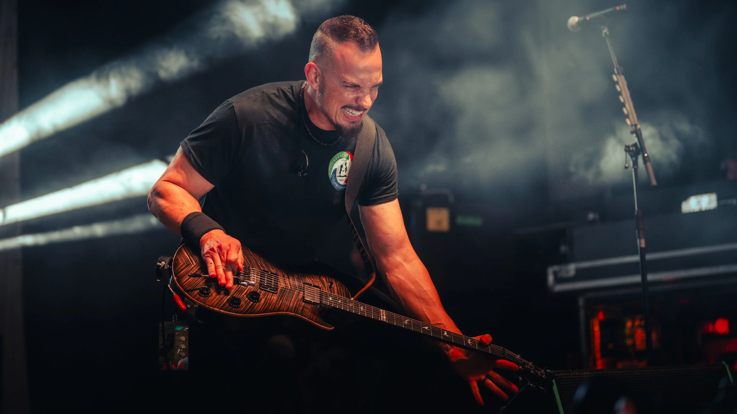 A male guitarist passionately performing on stage, with eyes closed and mouth open, holding a wood-colored electric guitar, in a smoky concert setting.
