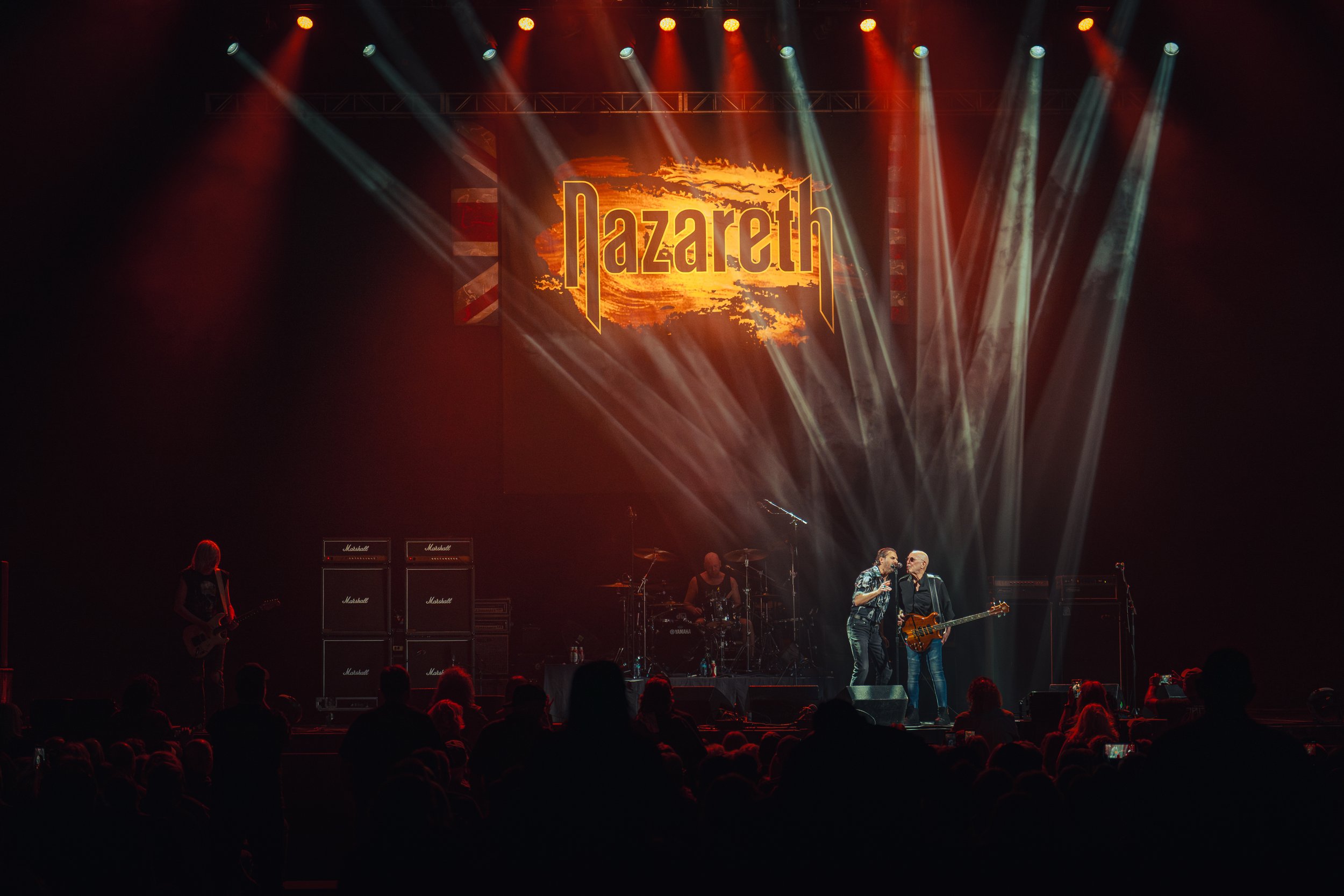 A band named Lazareth performing on stage with bright orange and white lights, a large screen displaying the band’s name, and an audience in the foreground.