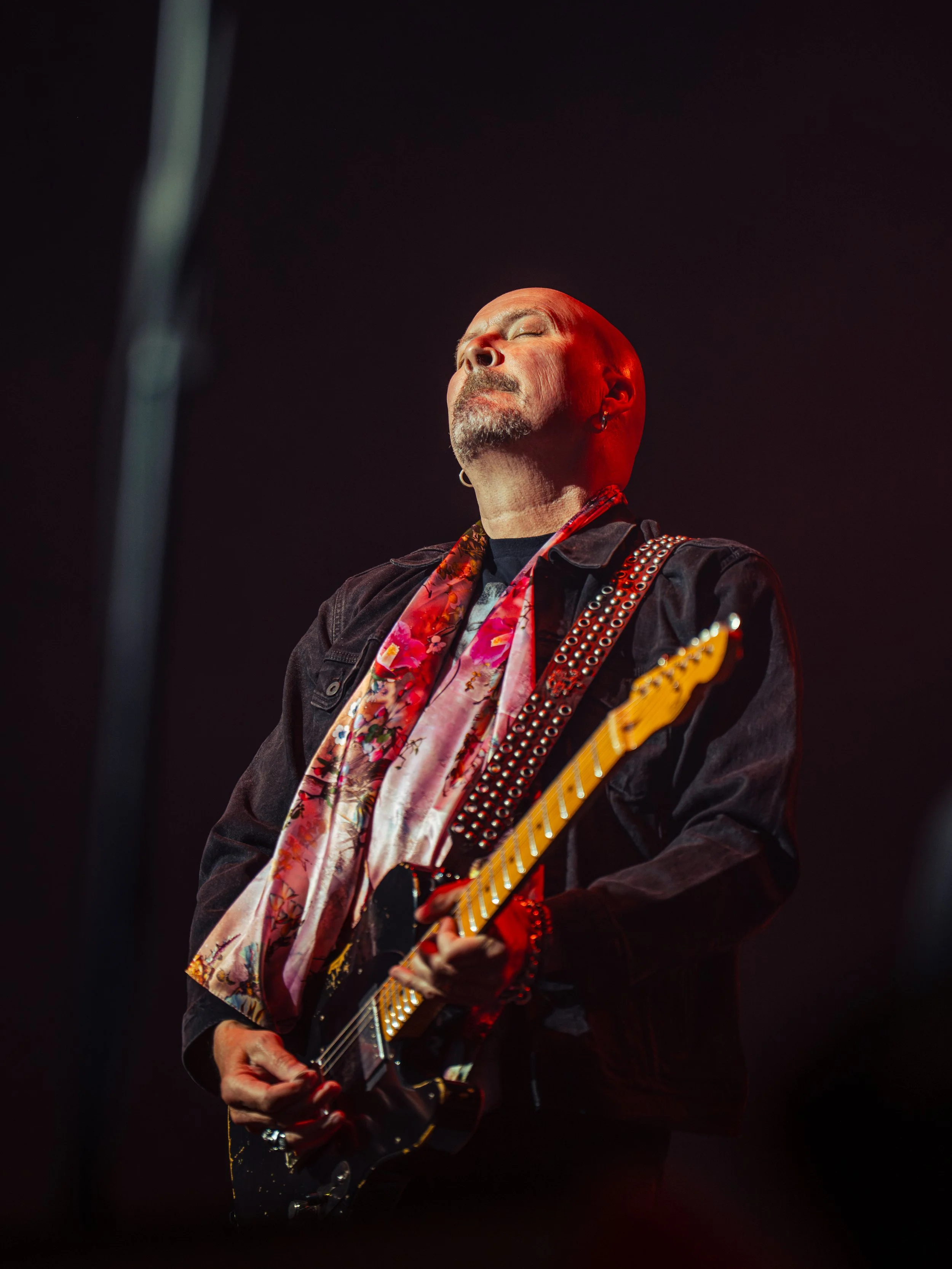 A man with a shaved head and beard playing an electric guitar on stage, wearing a dark jacket and a colorful scarf, with closed eyes and a serene expression.