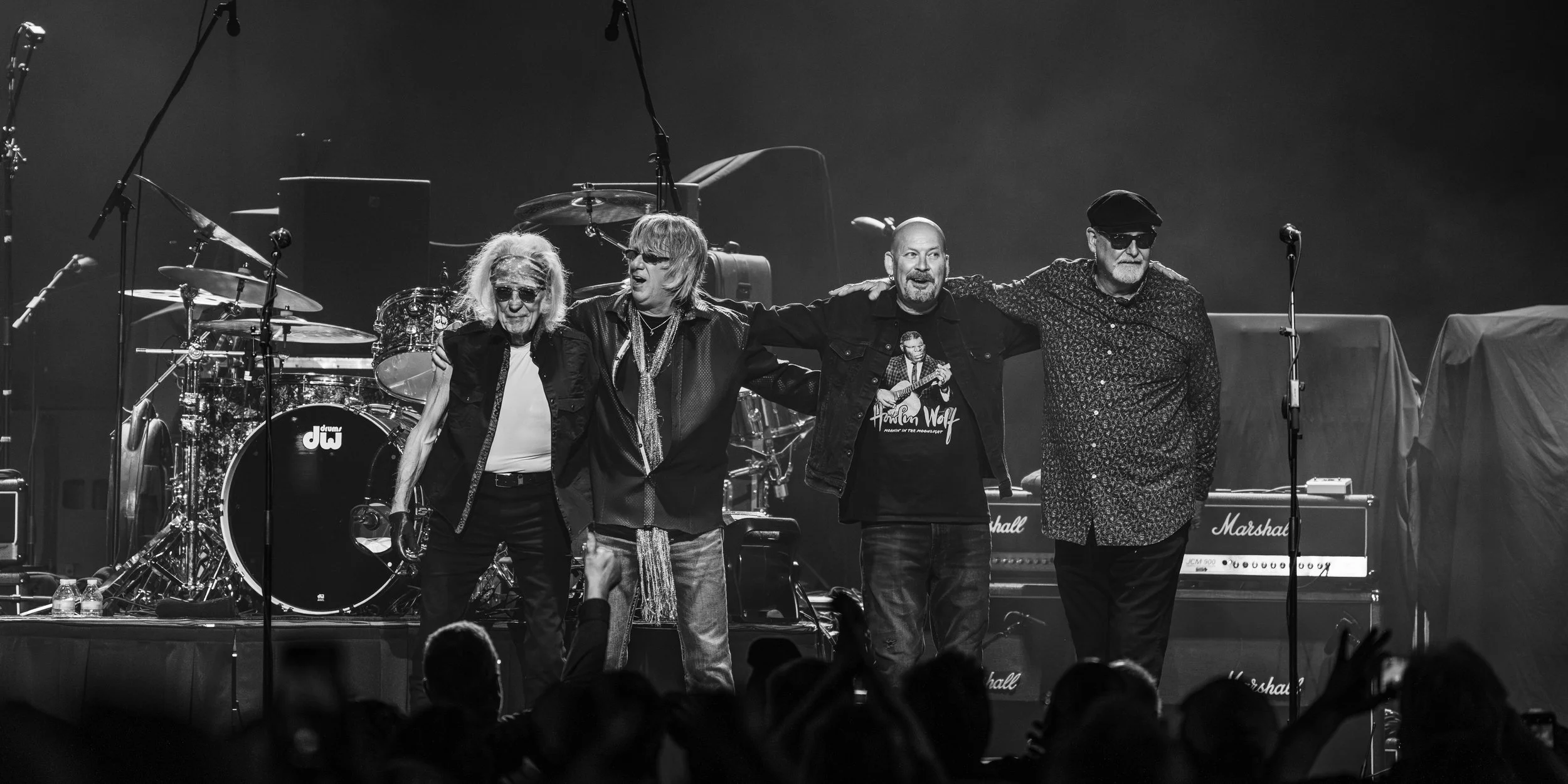 Four male musicians stand together on stage after a performance, arms around each other's shoulders, with musical instruments and equipment in the background.