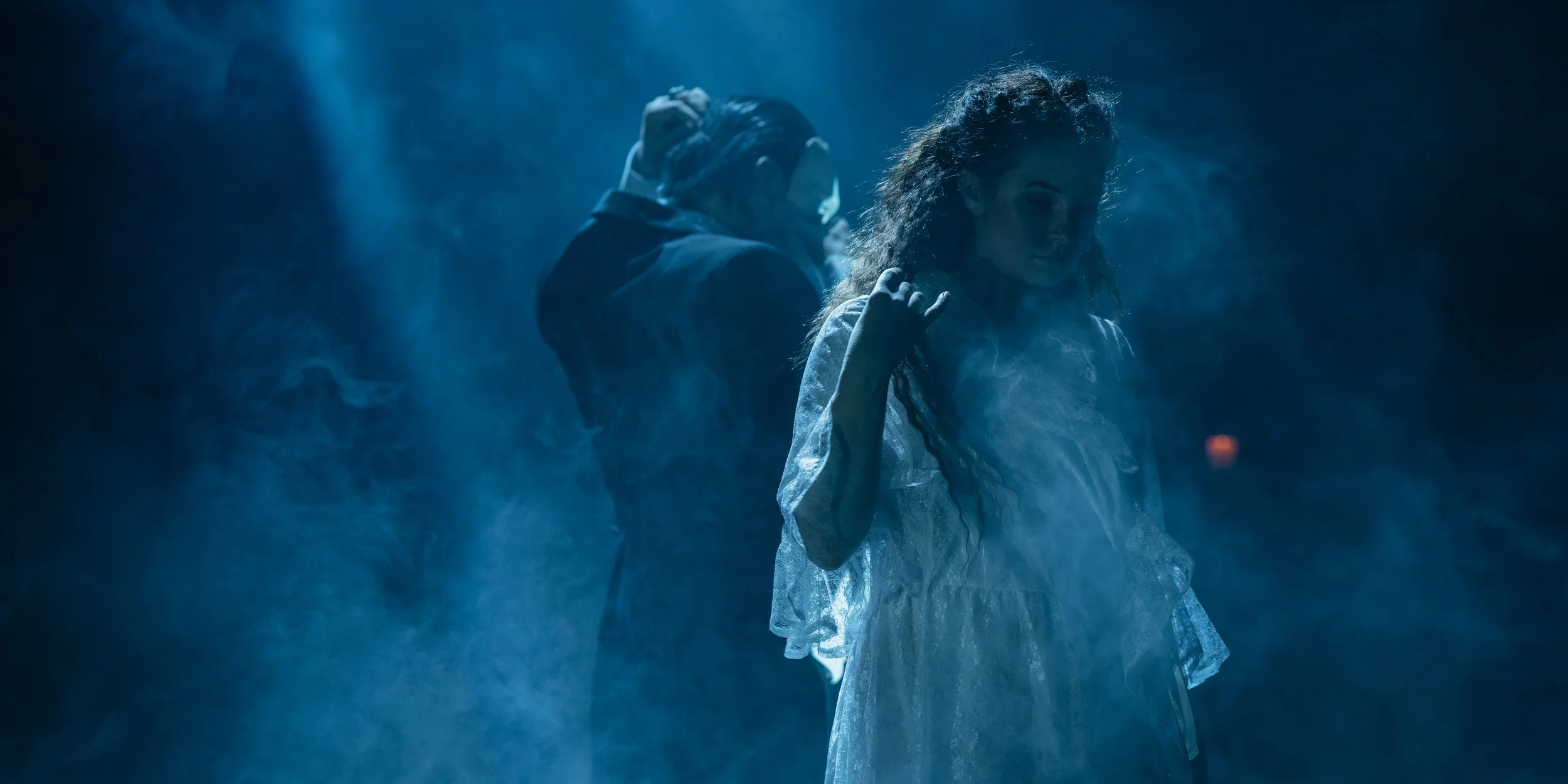 A woman in a historic or theatrical costume stands in a smoky, blue-lit environment with her head bowed and hand near her hair.