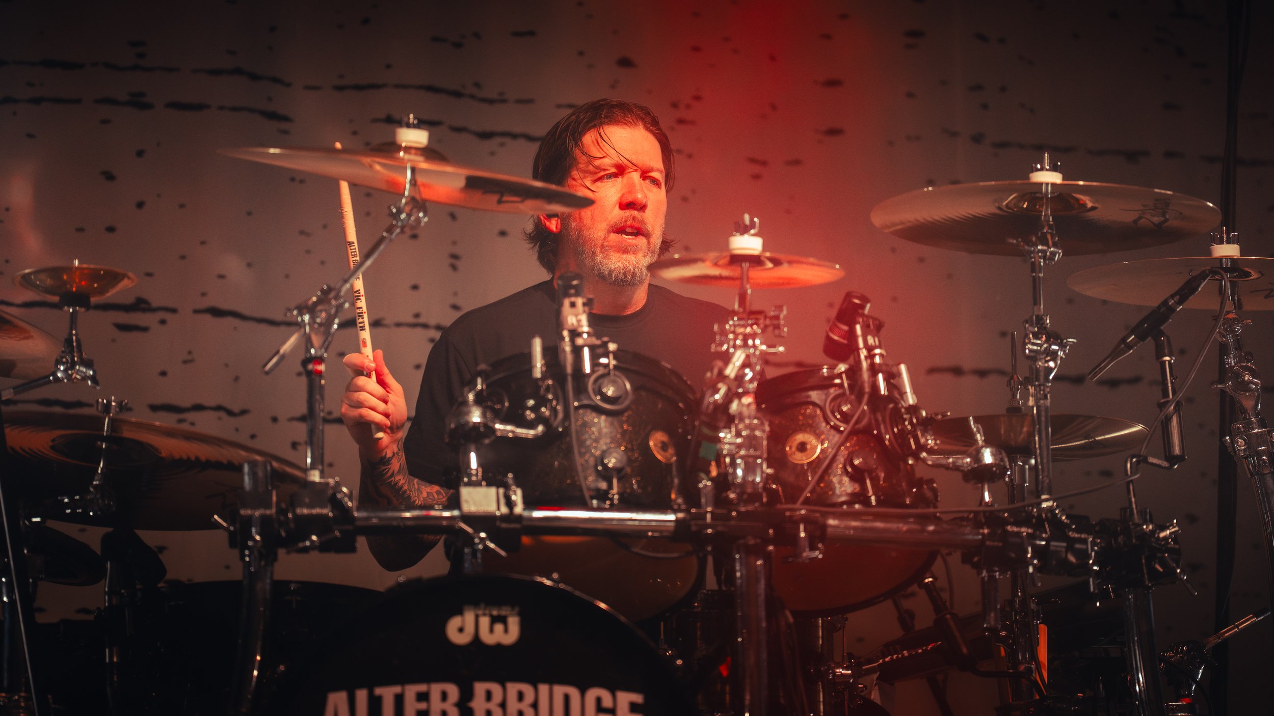 A male drummer with long hair and a beard playing a drum set on stage, with dim lighting and a dark abstract background.