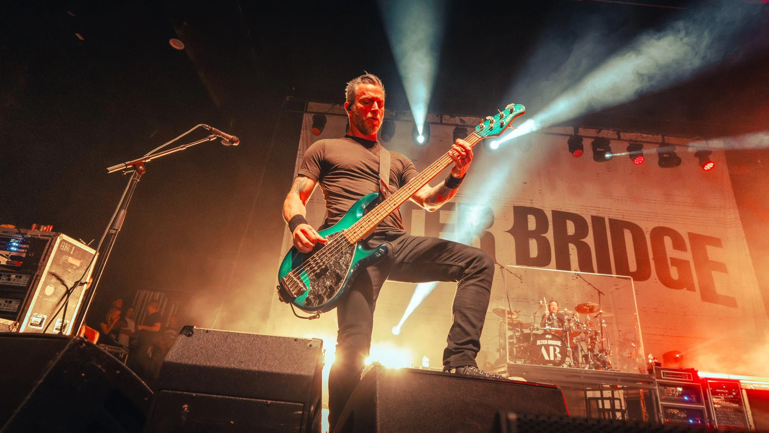 Musician playing an electric bass guitar on stage during a concert, with colorful stage lights and a drummer in the background.