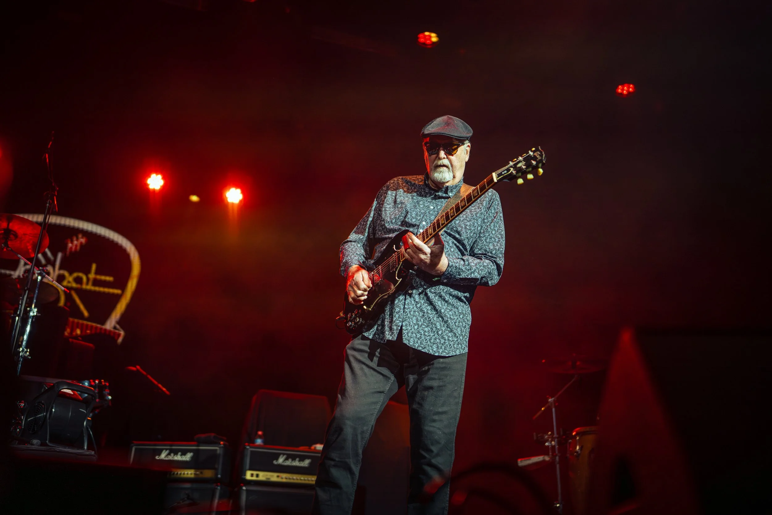 A man wearing sunglasses, a flat cap, and a patterned shirt playing an electric guitar on stage with red lighting and Marshall amplifier speakers in the background.