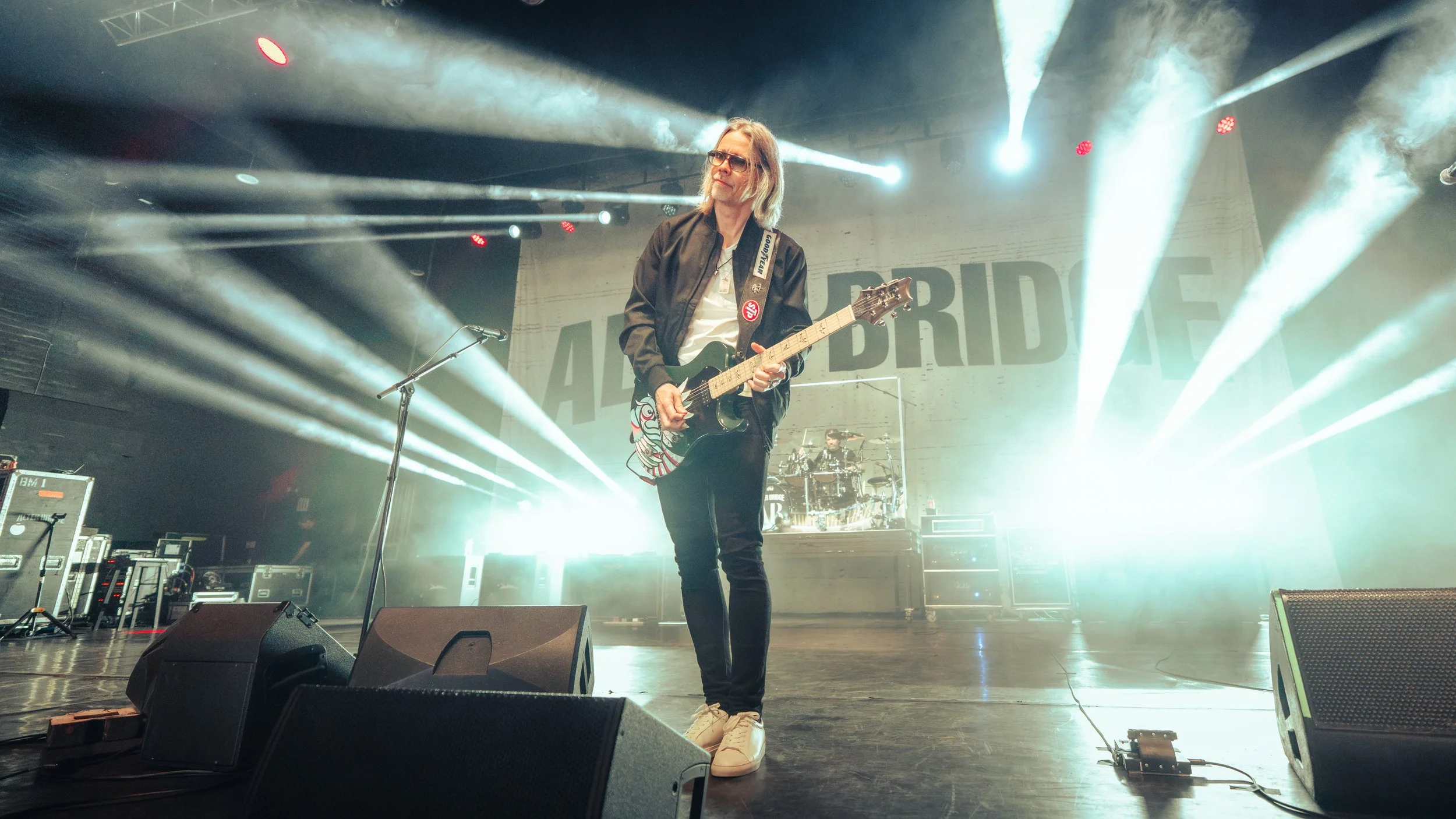 A musician with long hair and glasses playing an electric guitar on stage, surrounded by stage lights and fog, with a drummer in the background.