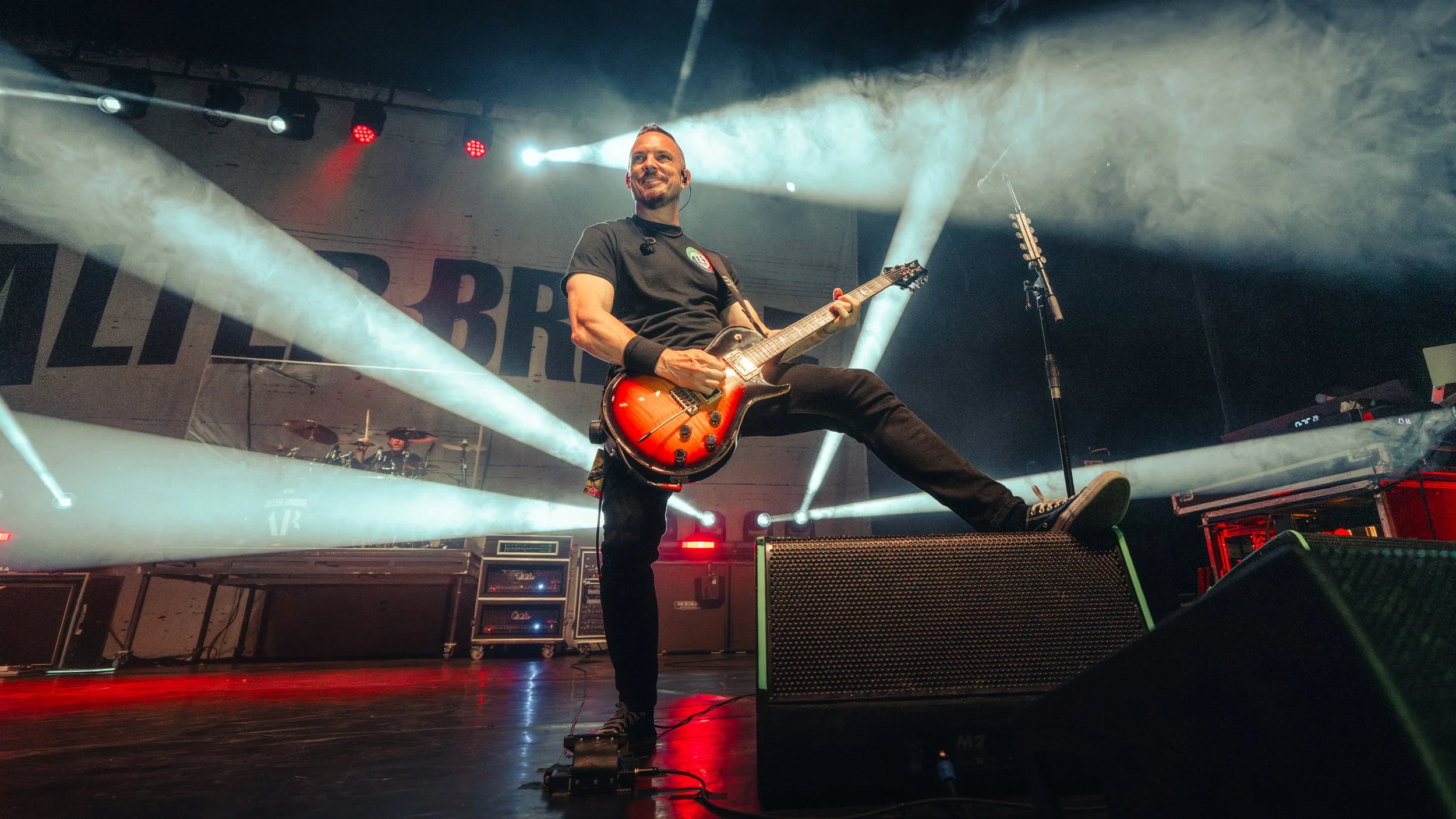 Male guitarist on stage at a concert, playing a red and black electric guitar, with stage lights and fog effects.
