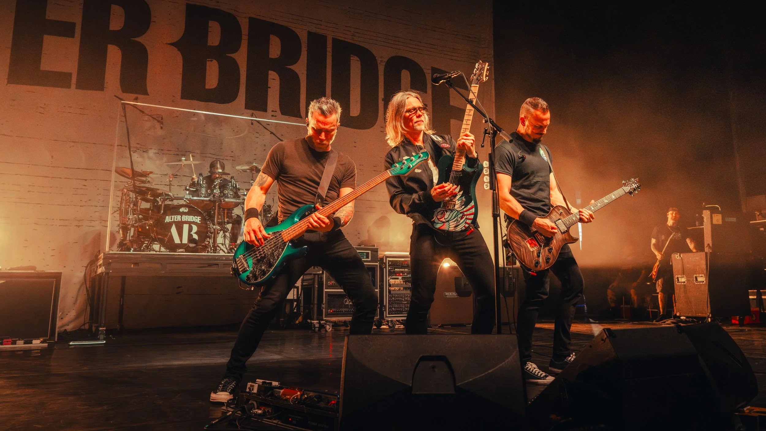 A band of three musicians performing on stage, one playing bass guitar, one singing into a microphone, and another playing electric guitar, with a drummer in the background, at a concert with a large sign reading 'ALTER BRIDGE' behind them.