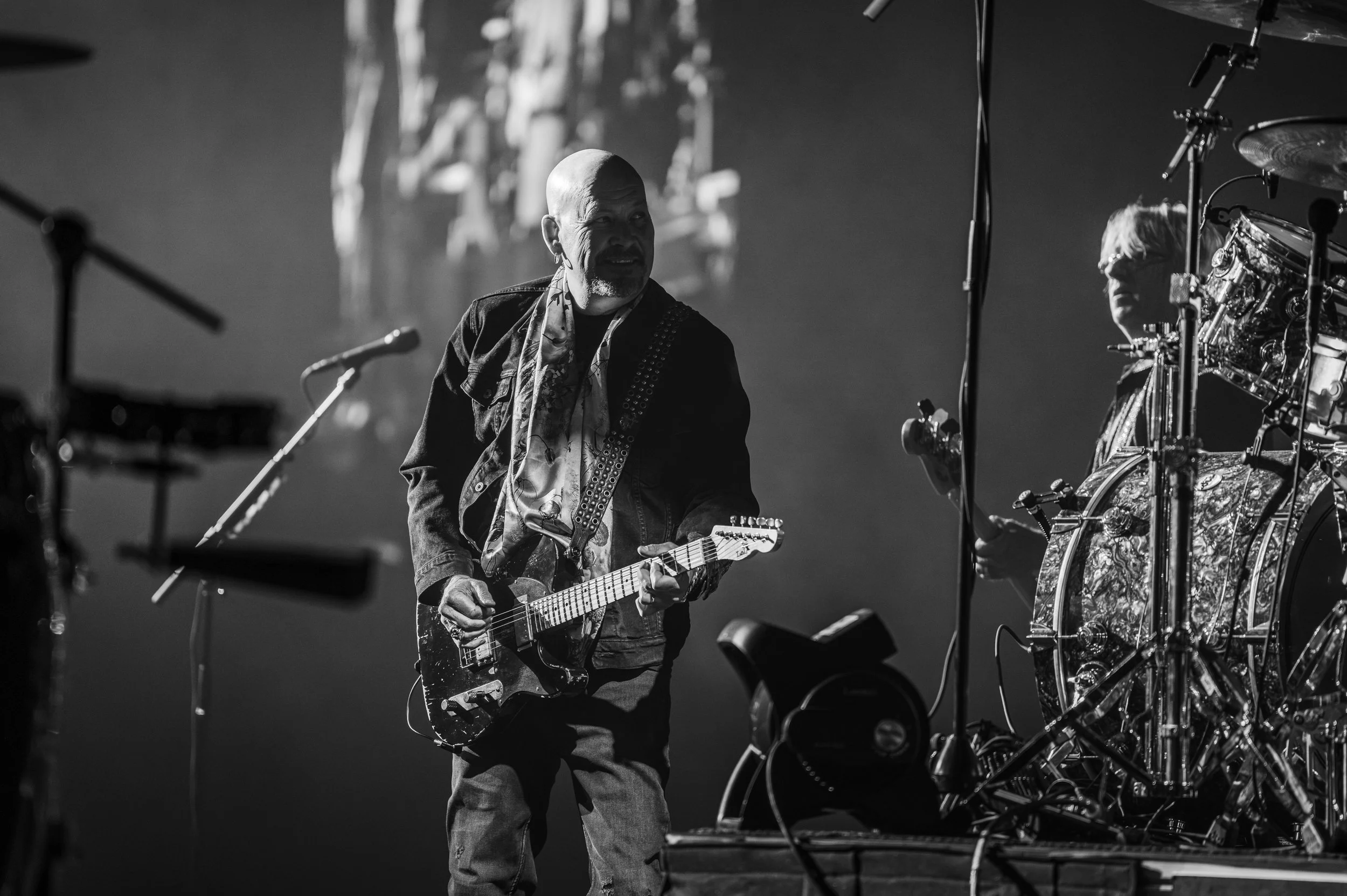A musician with a shaved head playing an electric guitar on stage, surrounded by musical equipment, with dramatic lighting in black and white.