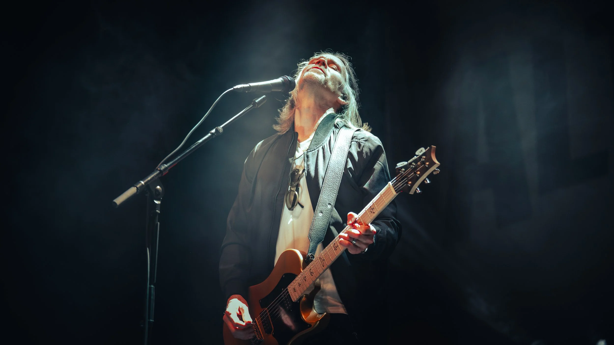 A musician performing on stage, singing into a microphone and playing an electric guitar, wearing a black jacket and glasses hanging from a necklace.