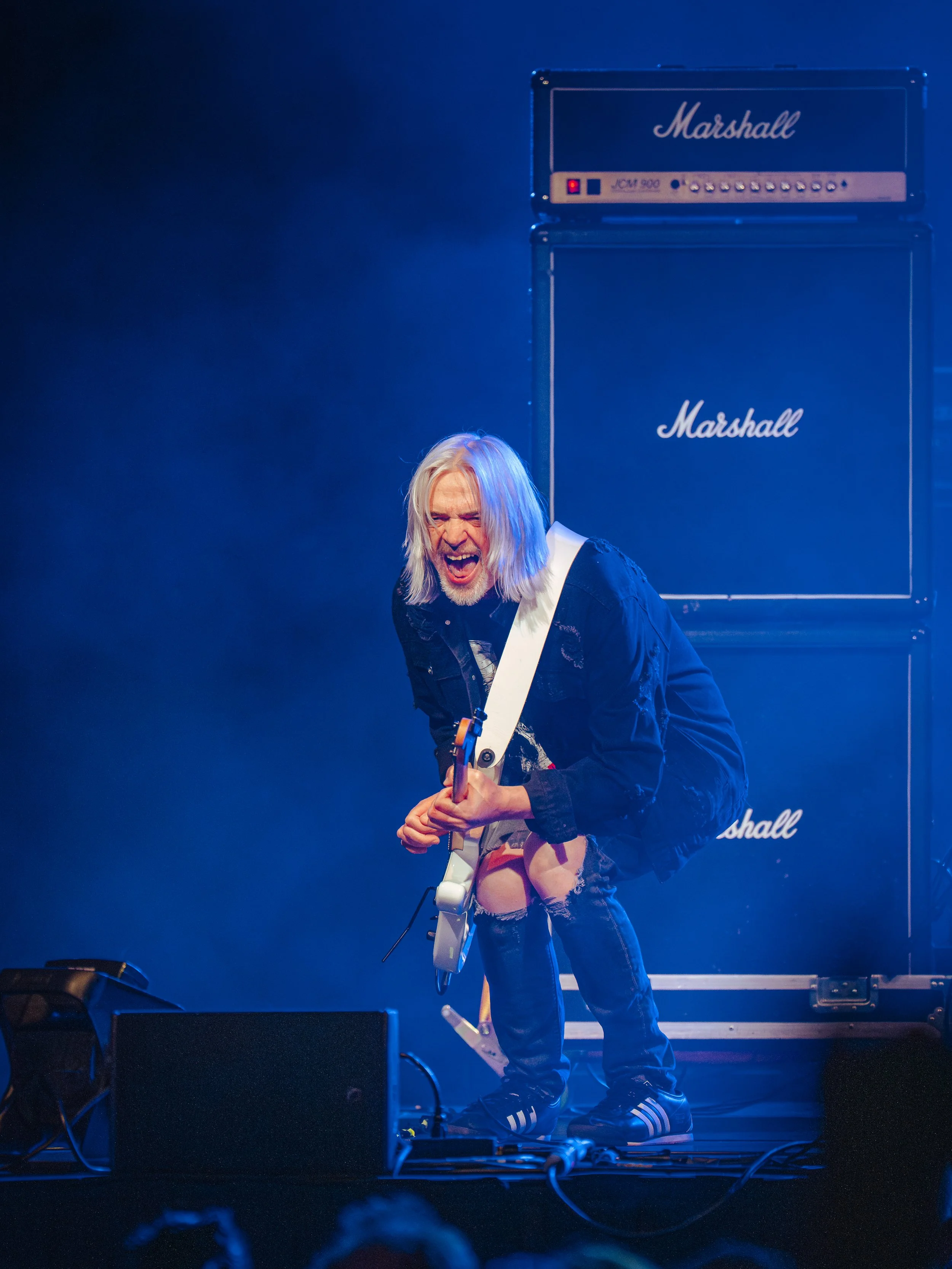Musician with long blonde hair playing an electric guitar on stage, with Marshall amplifiers in the background, under blue lighting.