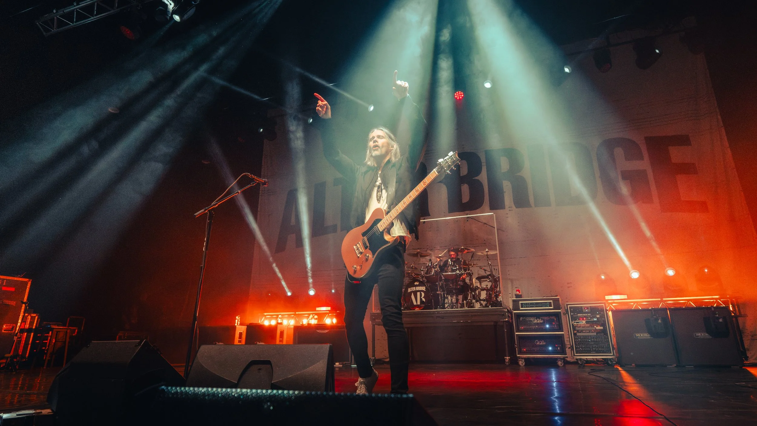 A musician with long hair playing an electric guitar on stage, with stage lights and band members visible in the background.
